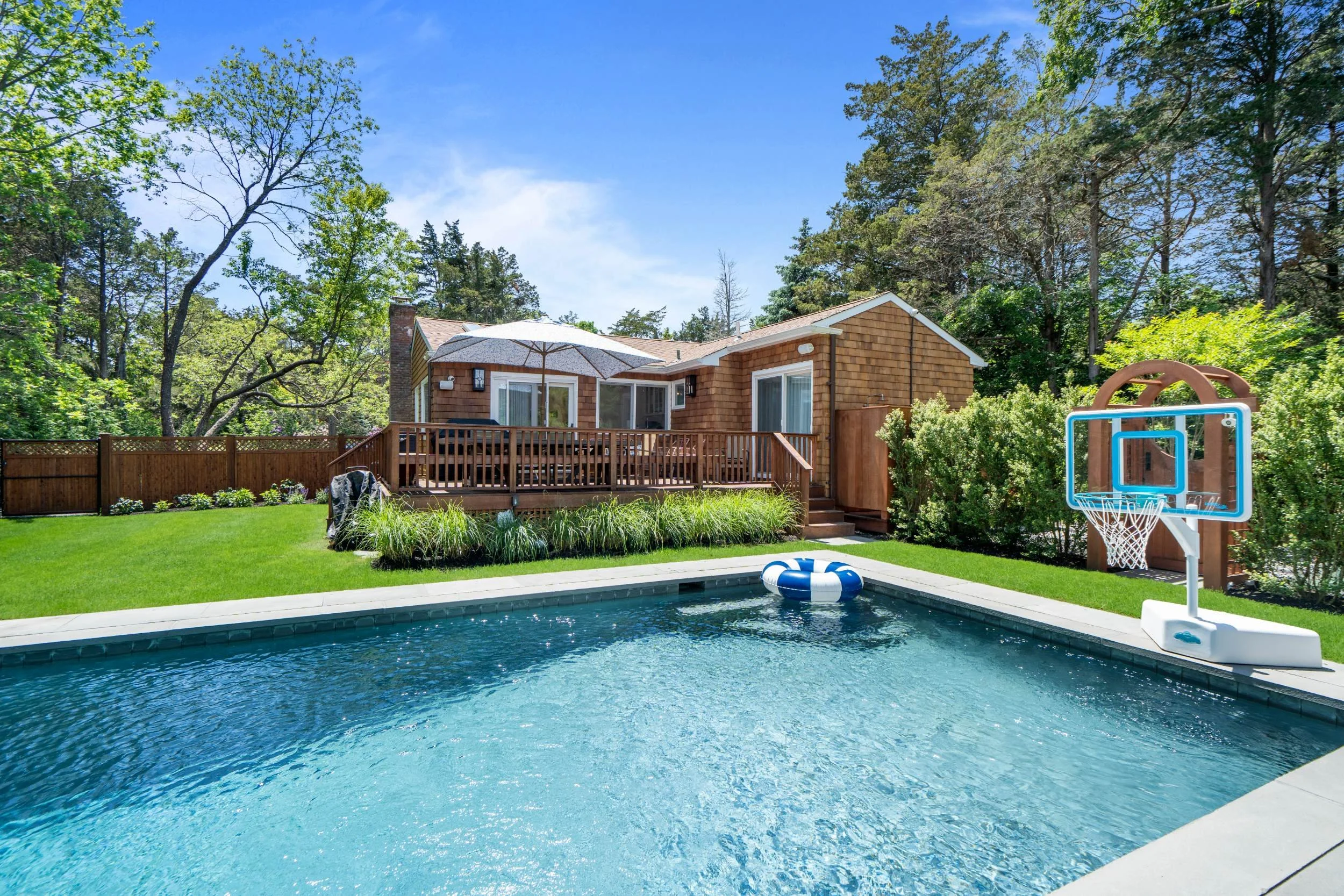 A backyard with a swimming pool, a wooden deck, and a house with large glass doors. The yard is surrounded by trees and greenery, and a basketball hoop stands near the pool.