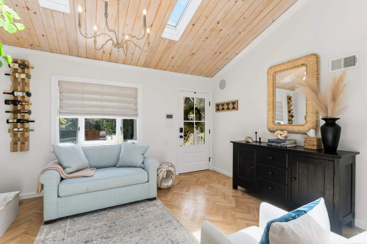 Living room with a light blue sofa, a black sideboard with decorative items, a large mirror, a wall-mounted wine rack, a window, and a skylight in the wooden ceiling.