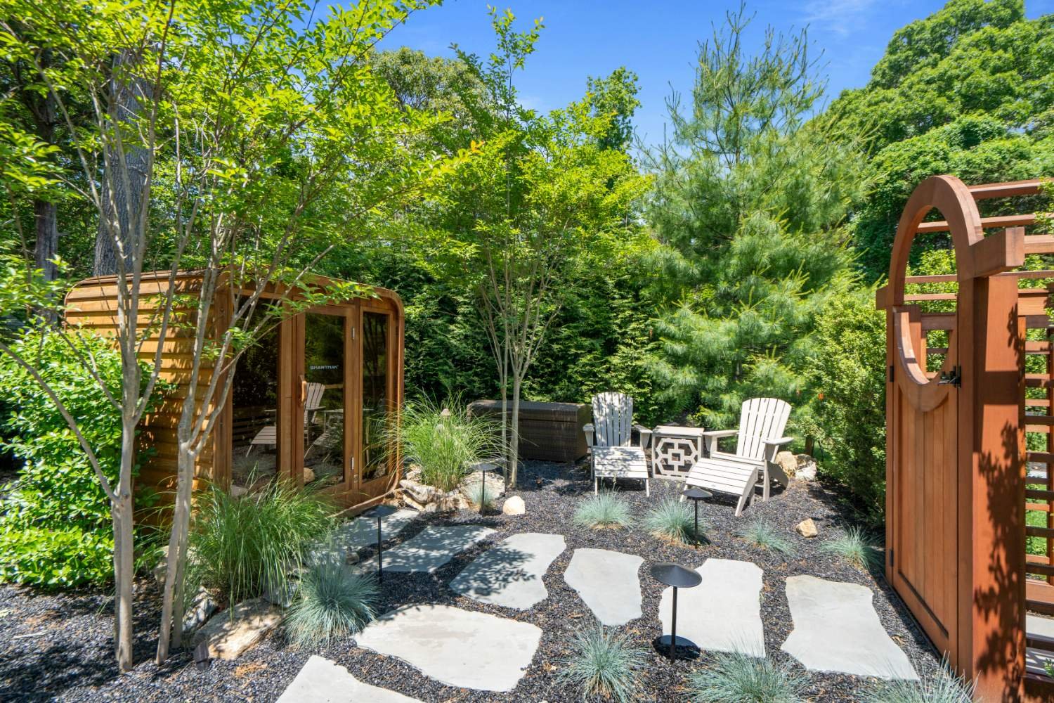 A cozy outdoor garden space surrounded by lush greenery, featuring white Adirondack chairs, a small table, and a wooden privacy screen with a circular cutout, with stone pathway and decorative plants.