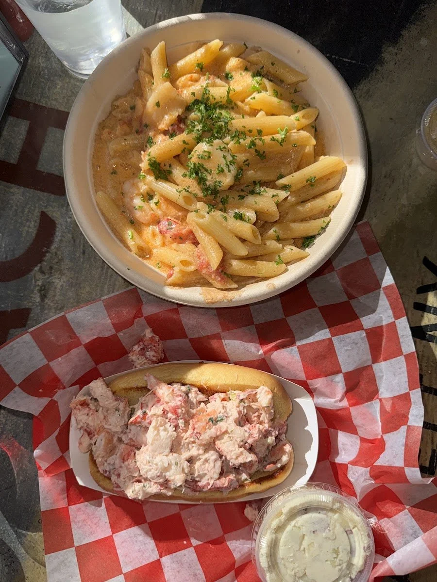 A bowl of shrimp pasta with a creamy sauce, garnished with chopped parsley. A lobster roll with lobster salad in a toasted bun, accompanied by a cup of soft serve ice cream.