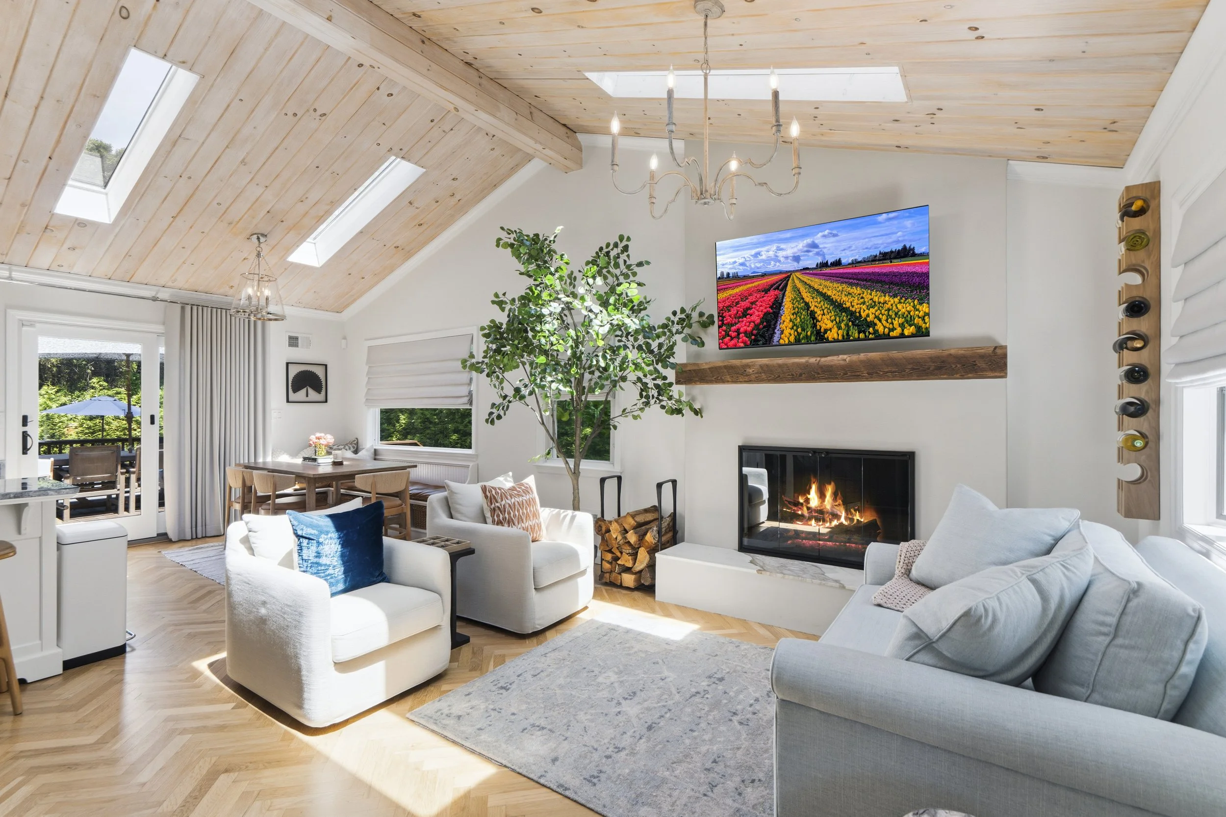 Living room with light wood vaulted ceiling, skylights, a chandelier, a large flat-screen TV showing a colorful tulip field, a fireplace, a gray couch, white armchair with a blue pillow, a wooden dining table with chairs, potted plant, and large wind