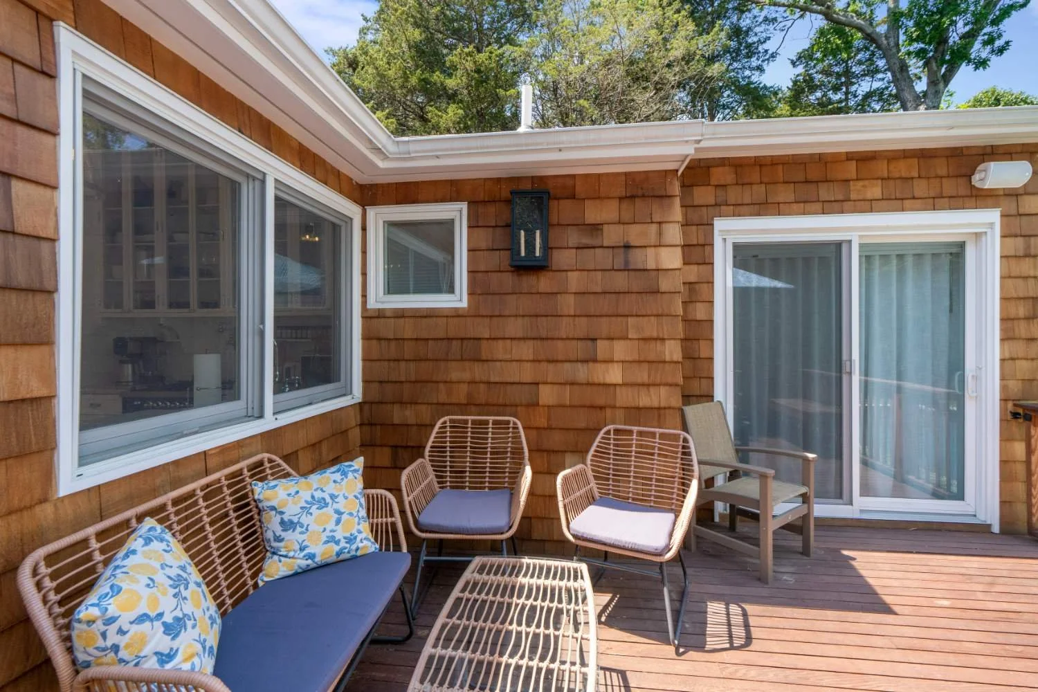 Outdoor wooden deck with wicker seating, cushions, and a sliding glass door leading inside a house with shingle siding, surrounded by green trees.