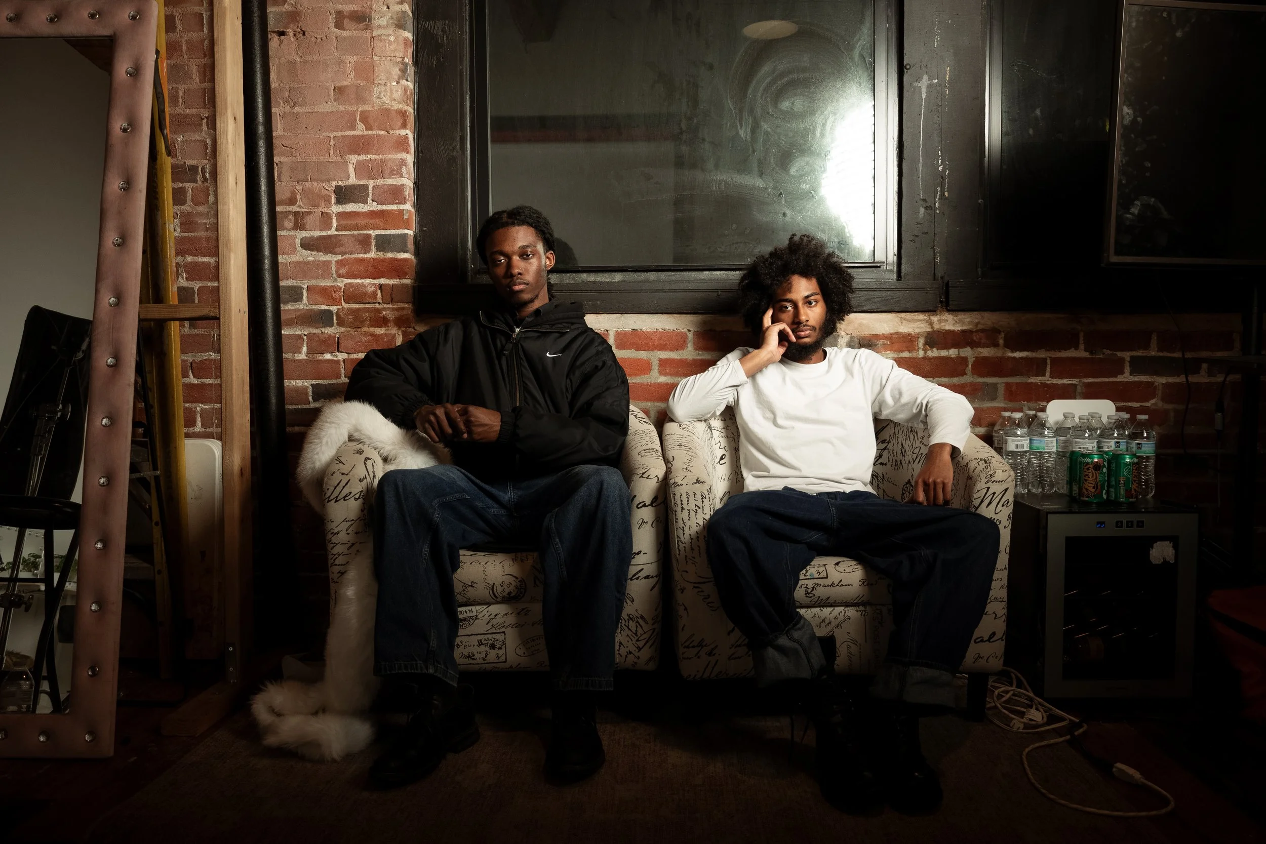 Two young men with dark skin sitting on a patterned sofa against a brick wall in an indoor setting. One is wearing a black Nike jacket and dark jeans, with his hands clasped. The other is wearing a white long-sleeve shirt and dark jeans, resting his 