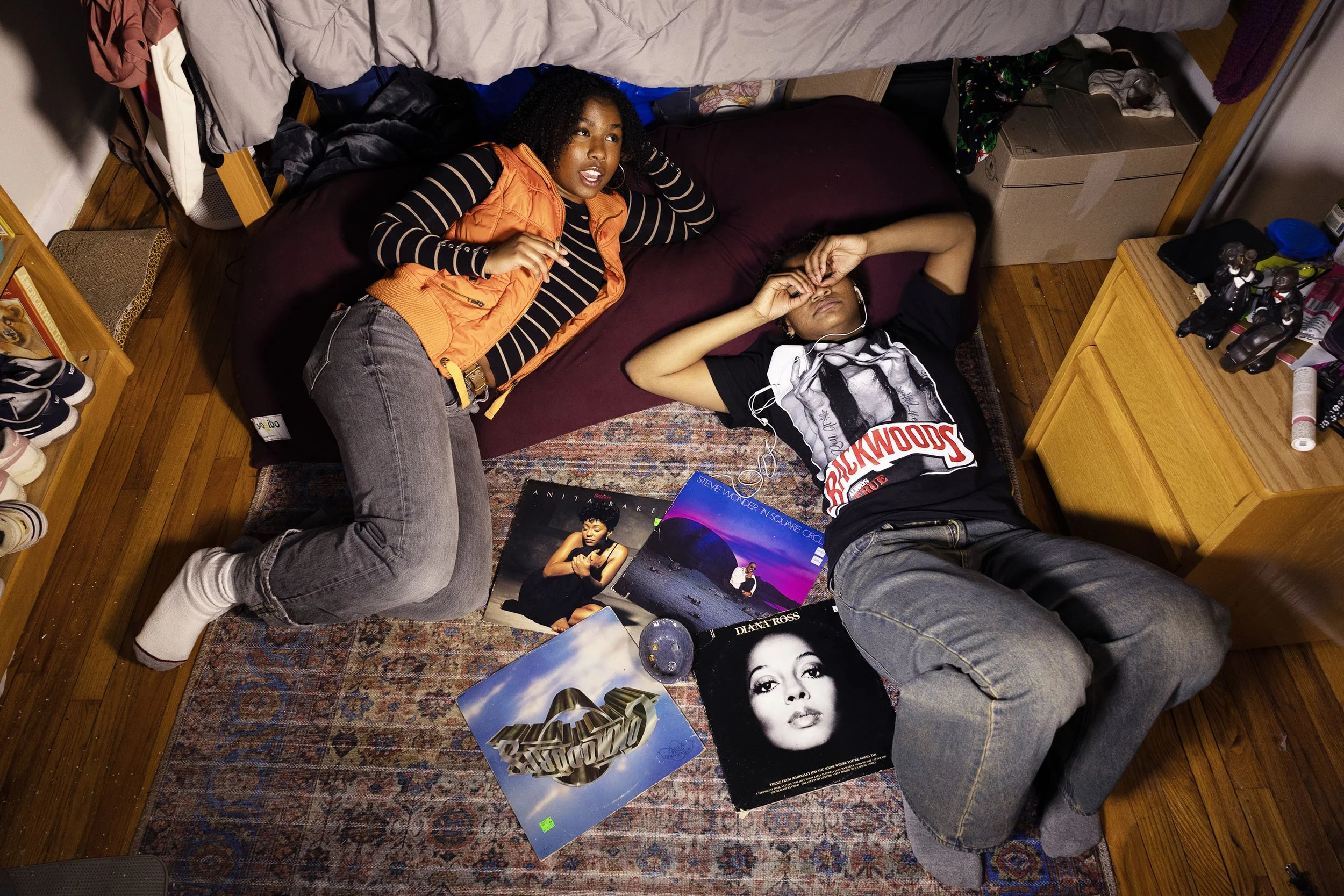 Two women are lying on a bed in a room. One woman is wearing an orange vest and striped long sleeve shirt, and the other woman is wearing a black and white graphic t-shirt. There are various magazines featuring a woman on the cover and a large gold h