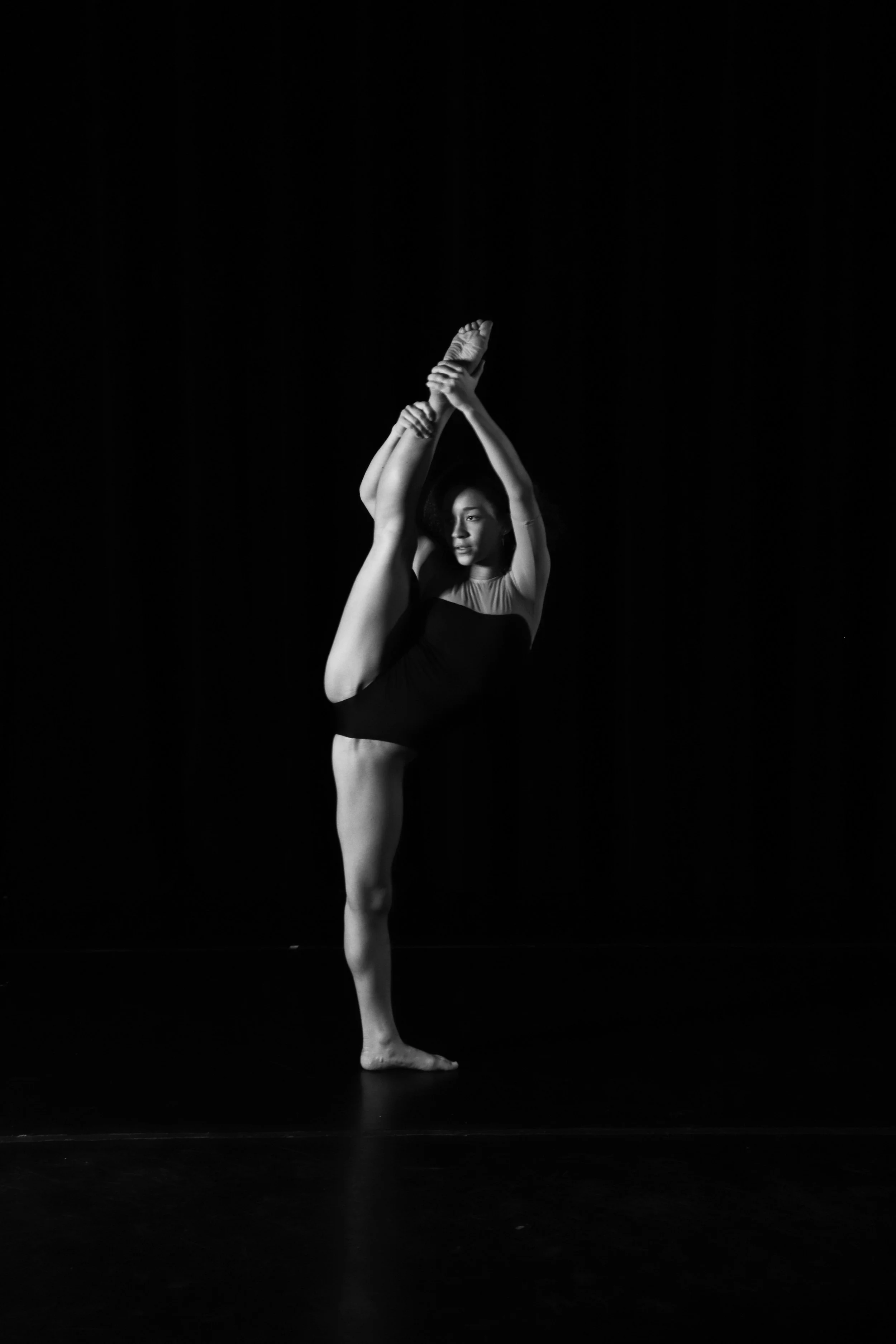 A dancer in a black outfit performs a standing split on a dark stage.