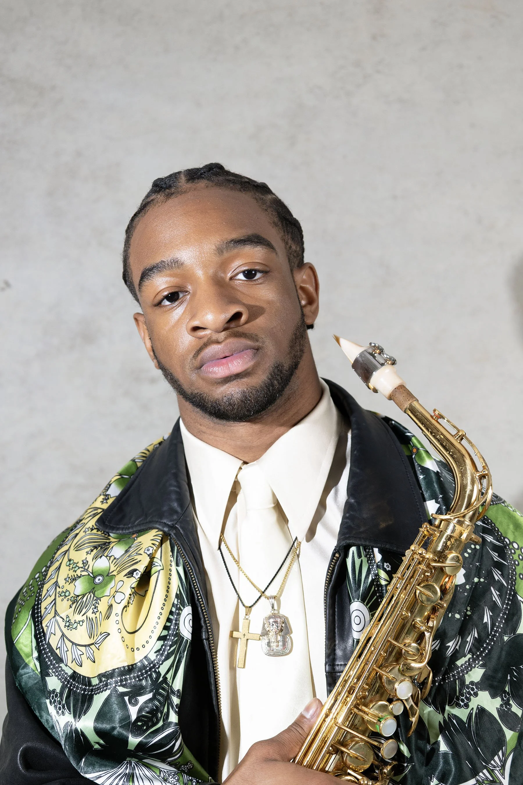 A young man with braided hair and a beard holding a gold saxophone, wearing a patterned jacket with black leather details and gold chains around his neck, posing against a plain grey background.