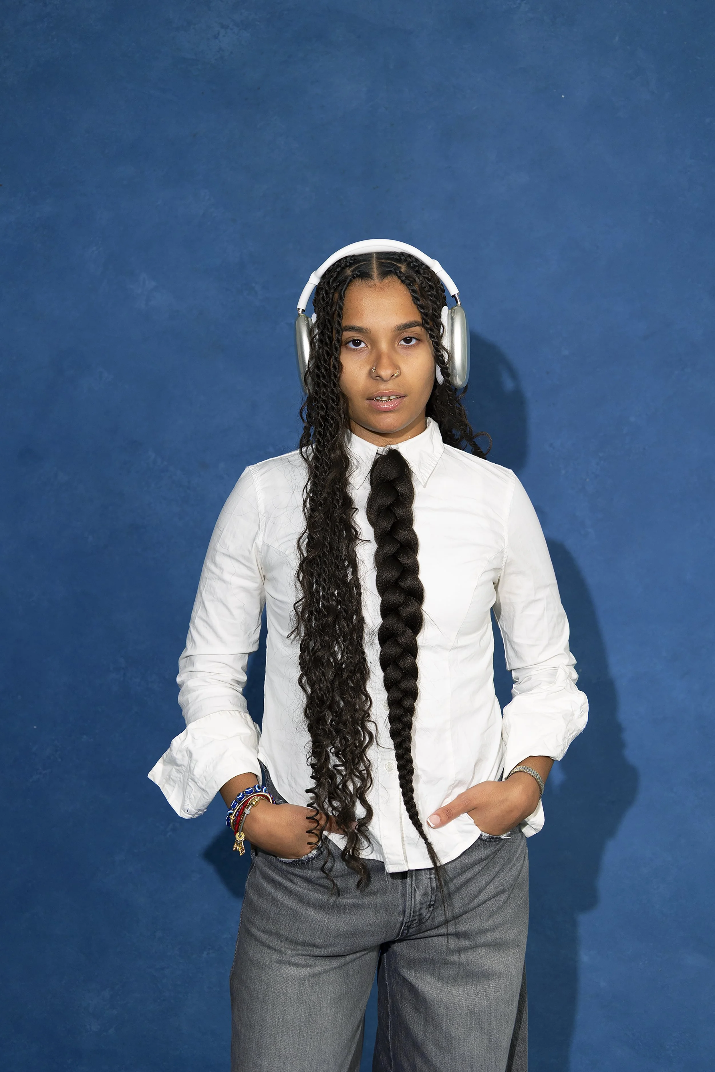 A young woman with long, curly hair wears a white button-up shirt, gray pants, multiple colorful bracelets, and headphones, standing against a blue background.