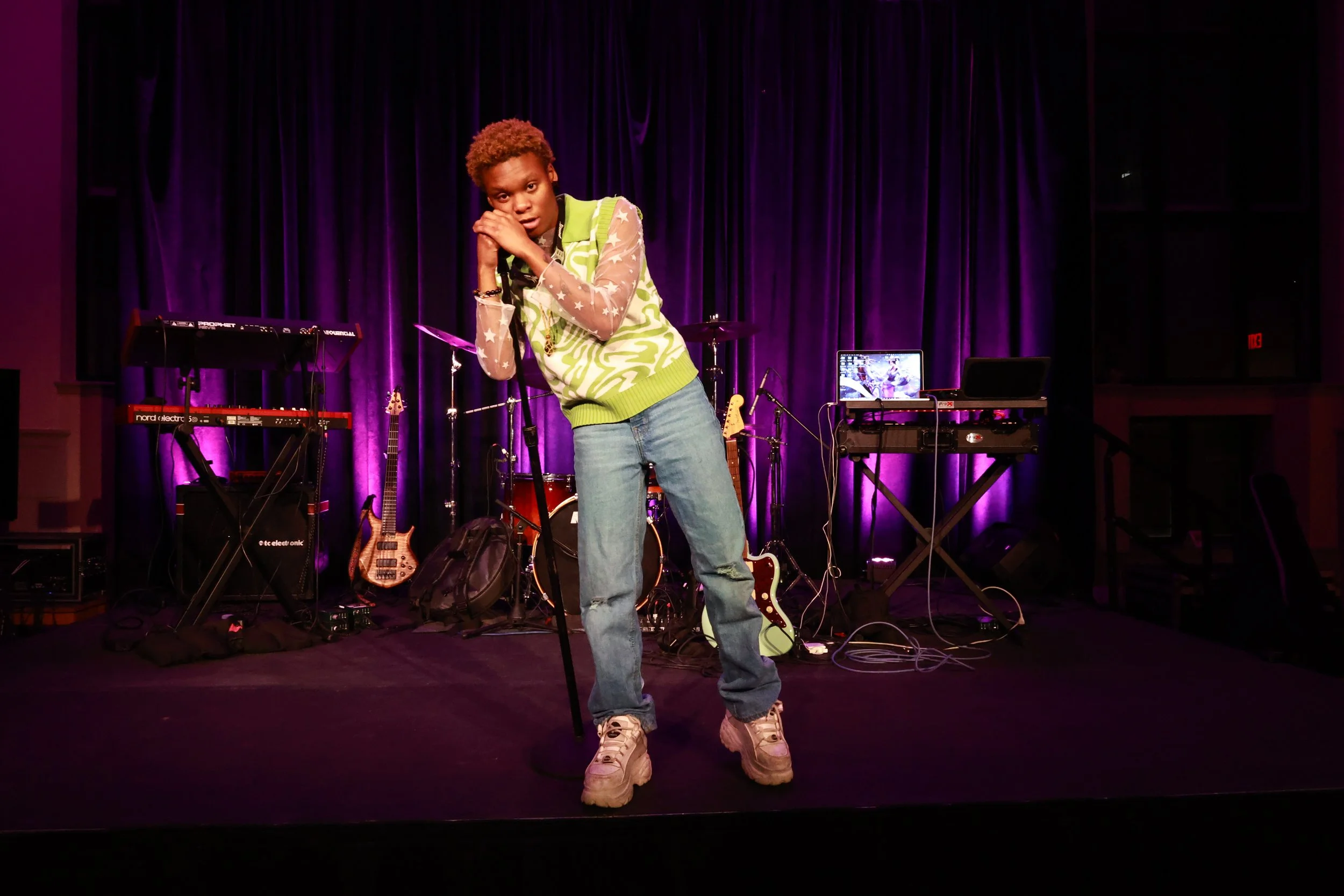 A young person with short curly hair standing on a stage with musical instruments and equipment, posing with hands together near their face, in front of a purple curtain.