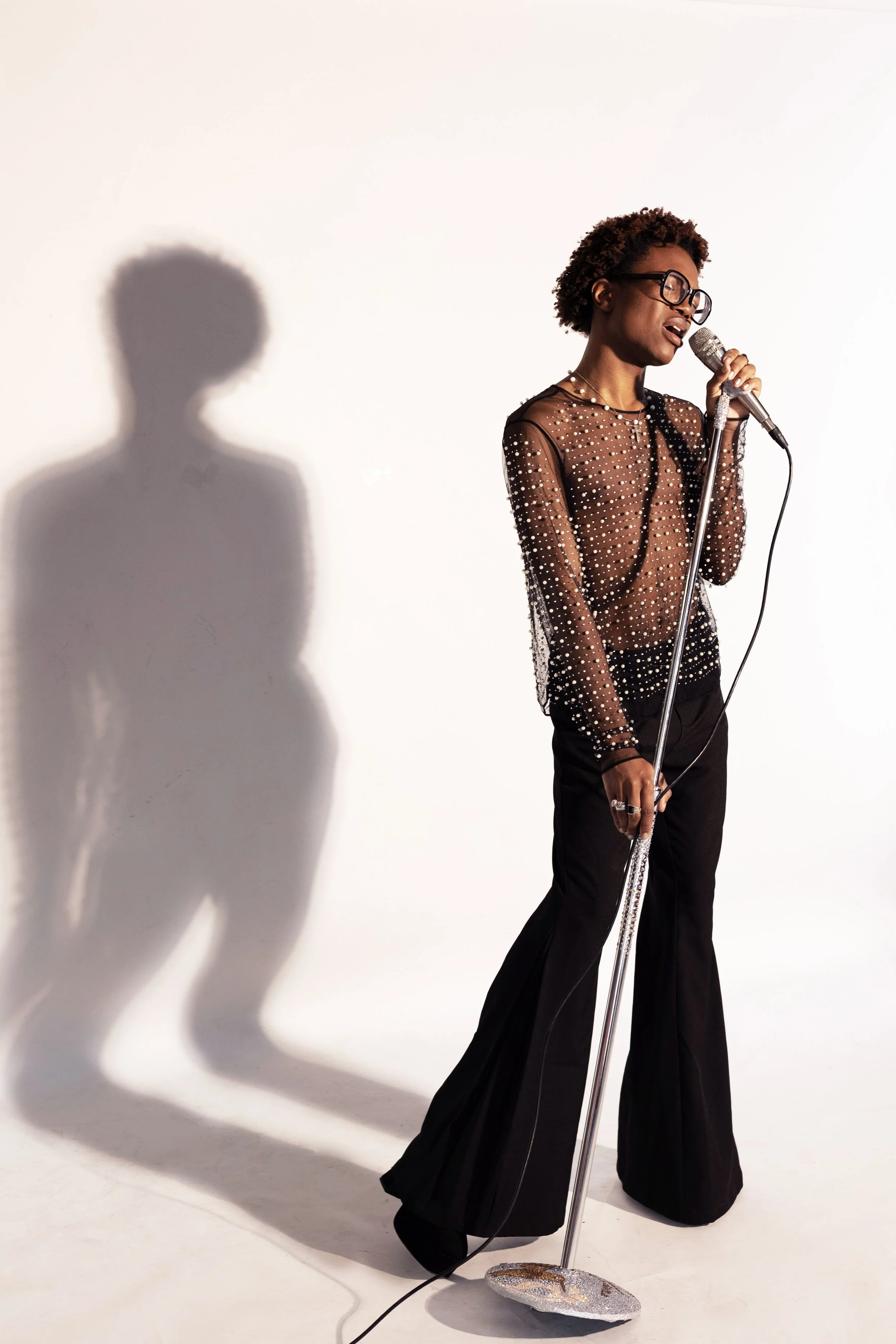 A woman with short curly hair, glasses, and a sheer black top with pearl embellishments, singing into a vintage-style microphone on a stand, with her shadow cast on a plain white wall.