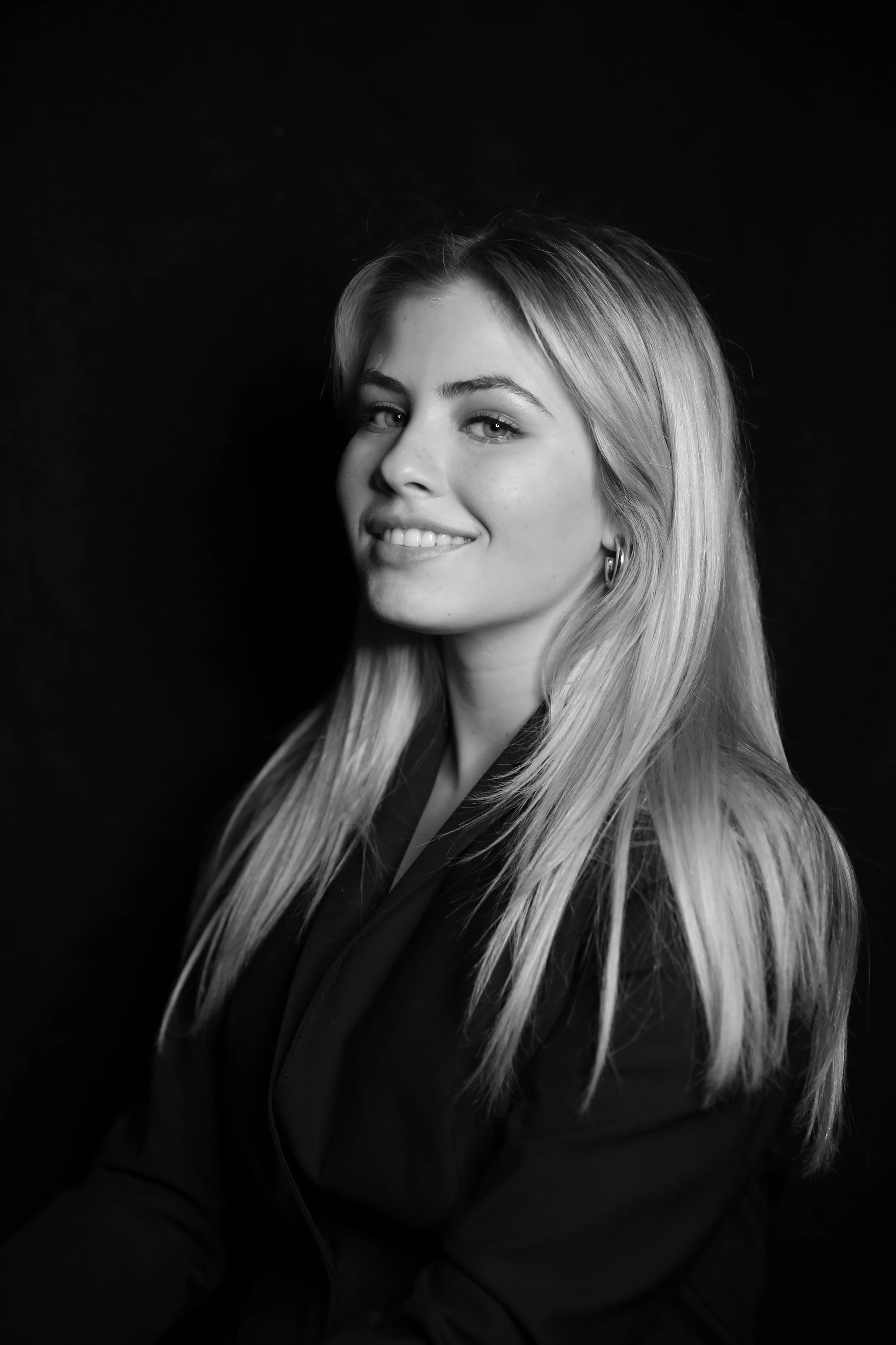 Black and white portrait of a young woman with long blonde hair, smiling slightly, wearing hoop earrings, against a dark background.