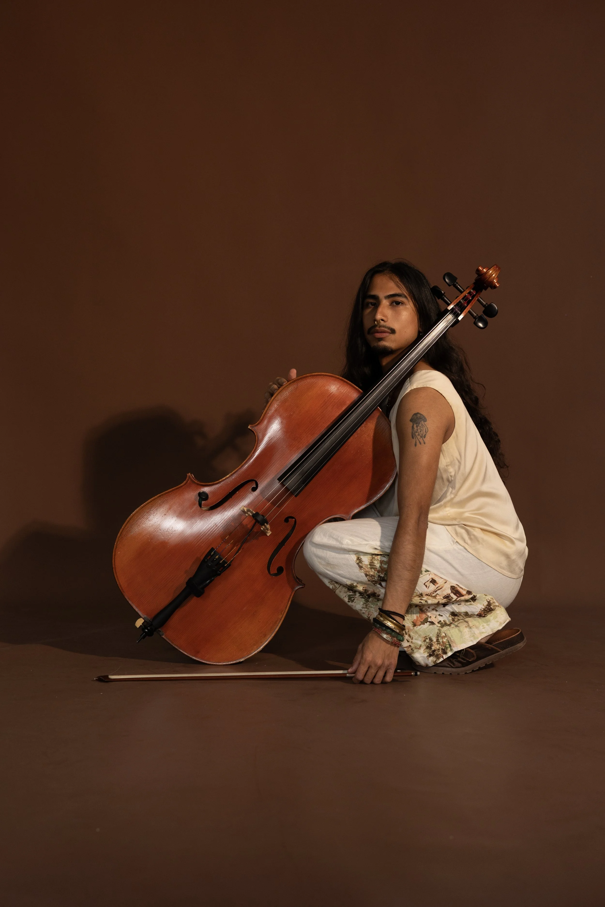 A person with long hair, tattoos, and piercings pose with a cello against a brown background. They are wearing a light sleeveless top and floral skirt and are kneeling on the ground.