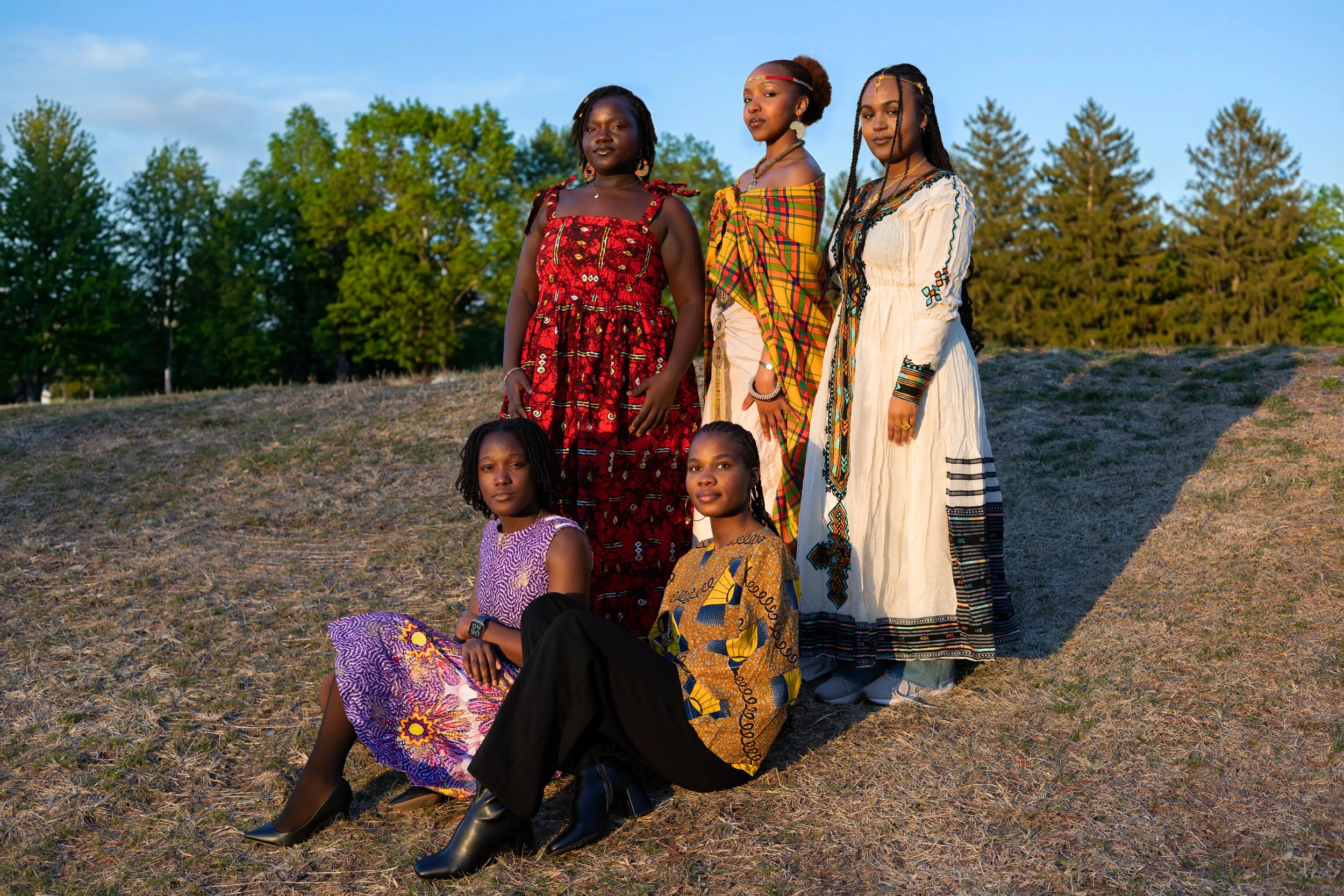 Group of four African women standing on a grassy hill with trees in the background, dressed in colorful traditional attire, with two women seated in front.