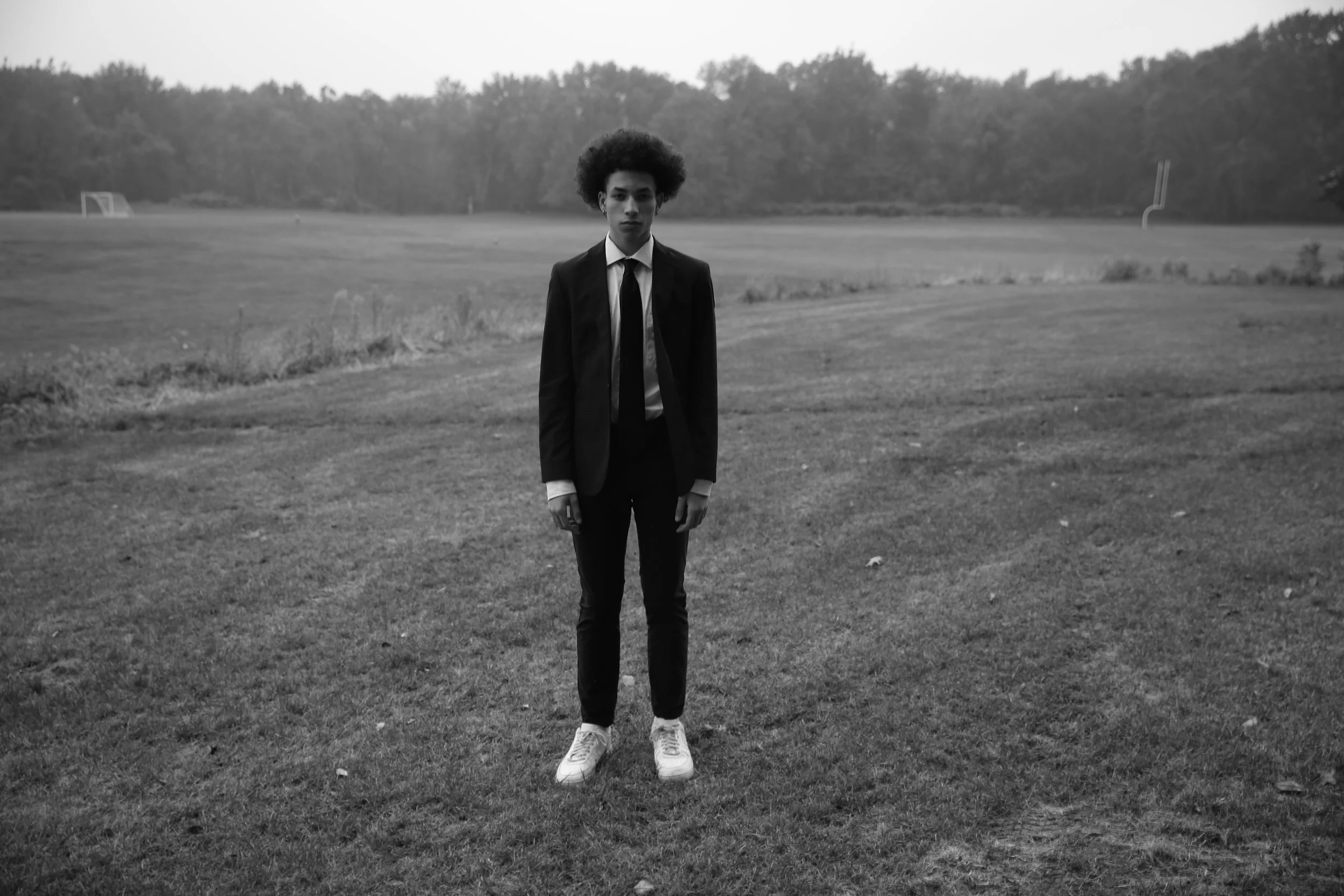 A young man is standing alone in the middle of a grassy field, wearing a suit and tie, with white sneakers. The field is open with trees in the background, and the sky is overcast.