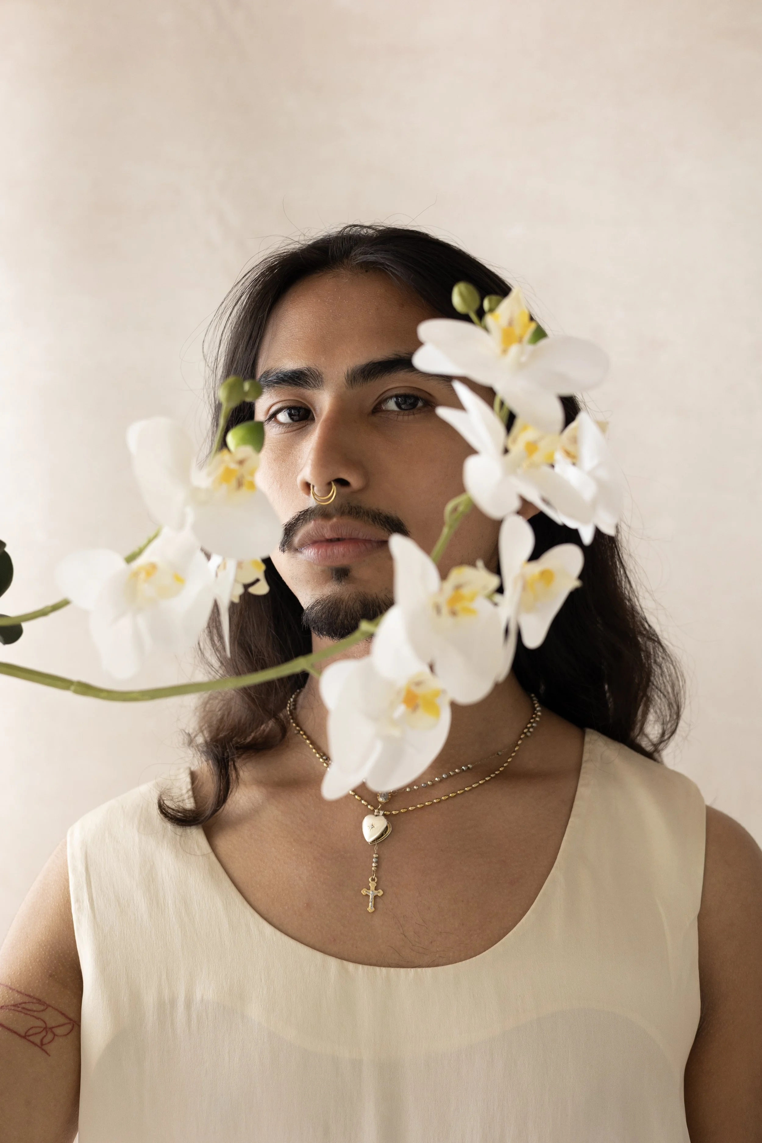 A woman with dark hair and a septum piercing holding white orchids in front of her face, wearing a beige sleeveless top, layered gold necklaces with a cross and heart pendant, standing against a plain neutral background.