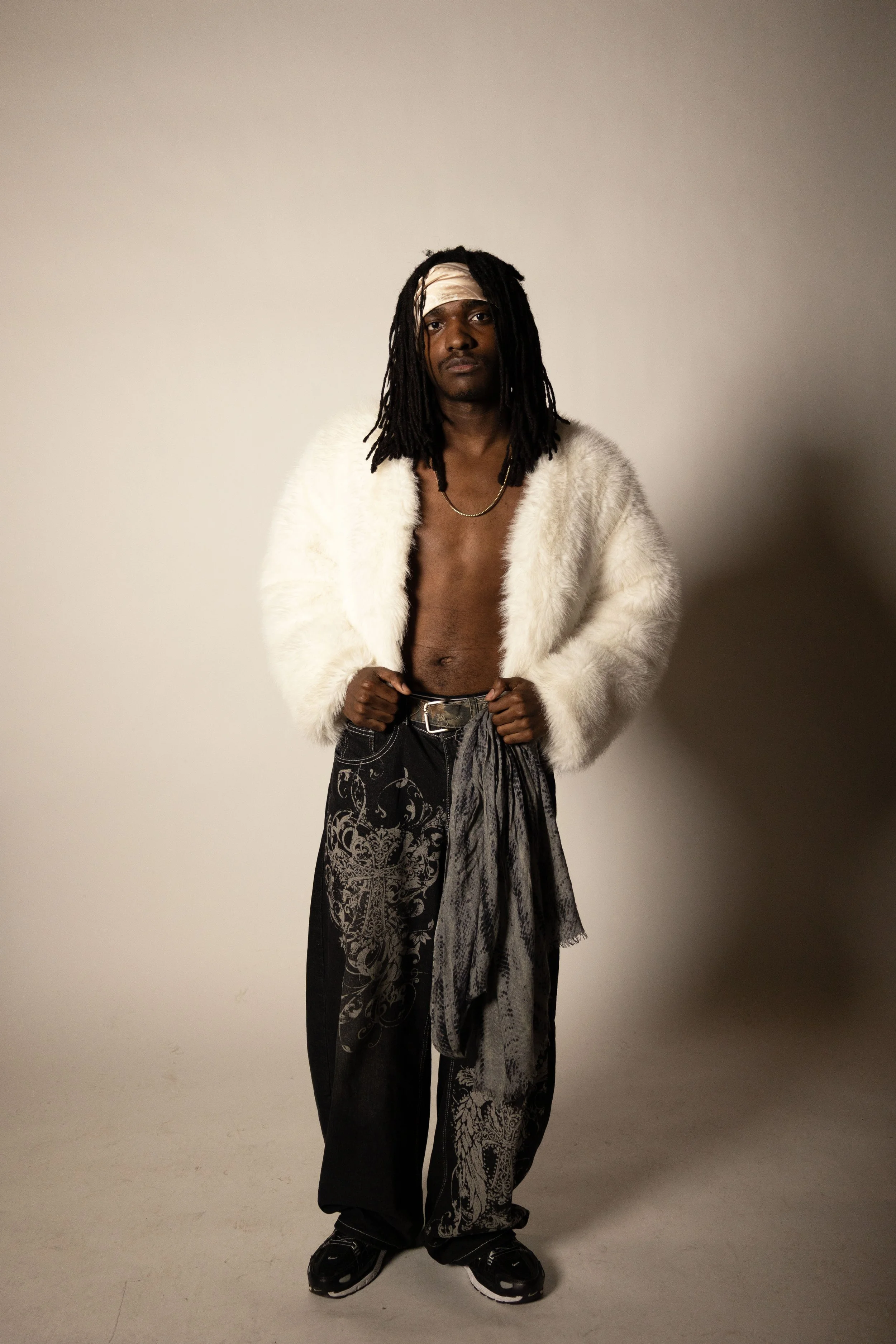 A man with long dreadlocks wearing a white faux fur jacket, black and gray patterned pants, sneakers, a light pink headband, and a gold chain necklace, standing against a plain background.