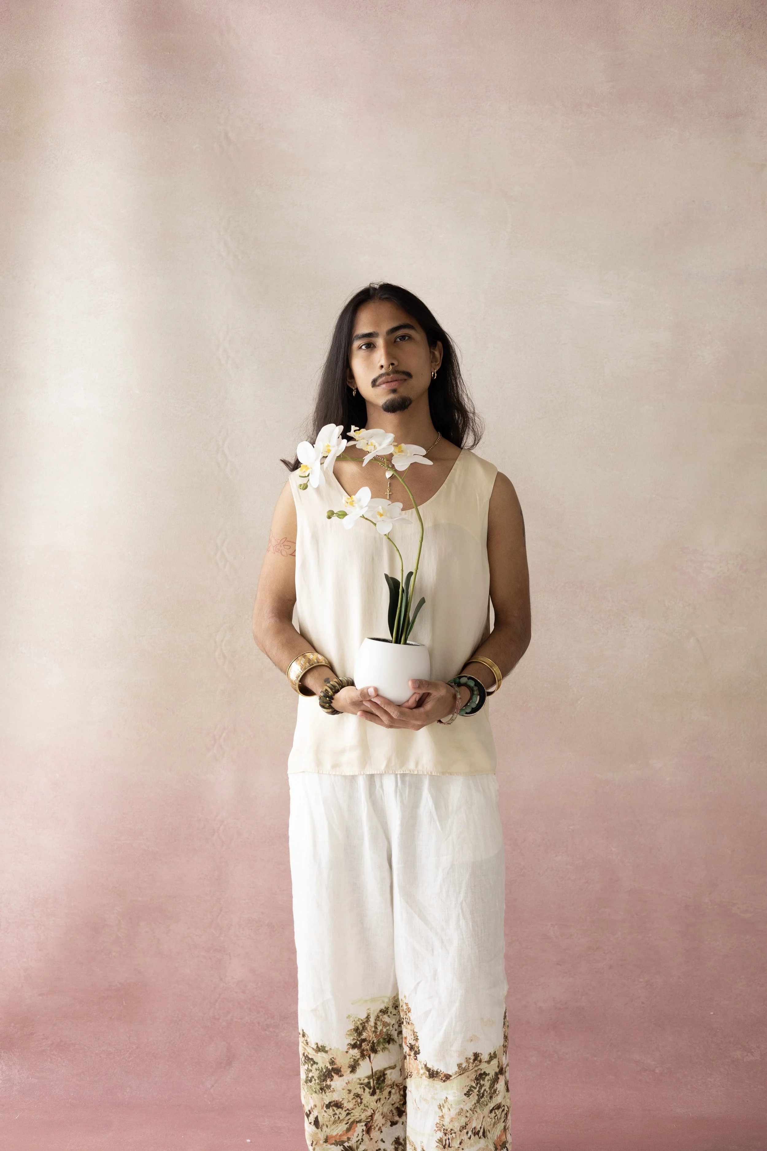 A person with long dark hair standing against a pinkish wall, holding a white vase with white orchids, wearing a cream sleeveless top and white pants with a landscape print at the bottom, accessorized with bracelets and earrings.