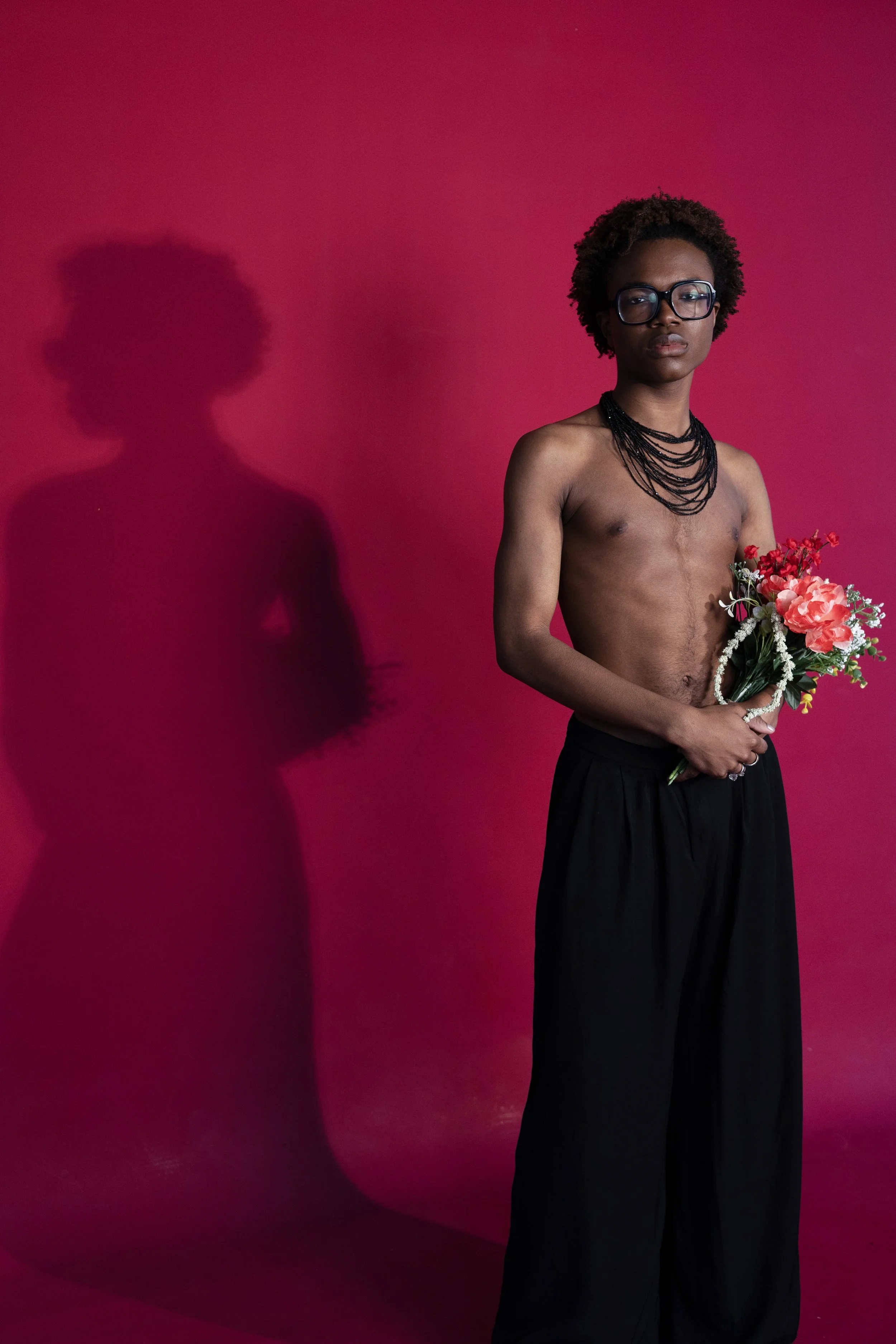 A topless woman with short curly hair and glasses holding a bouquet of flowers against a hot pink background. Her shadow is cast on the wall behind her.