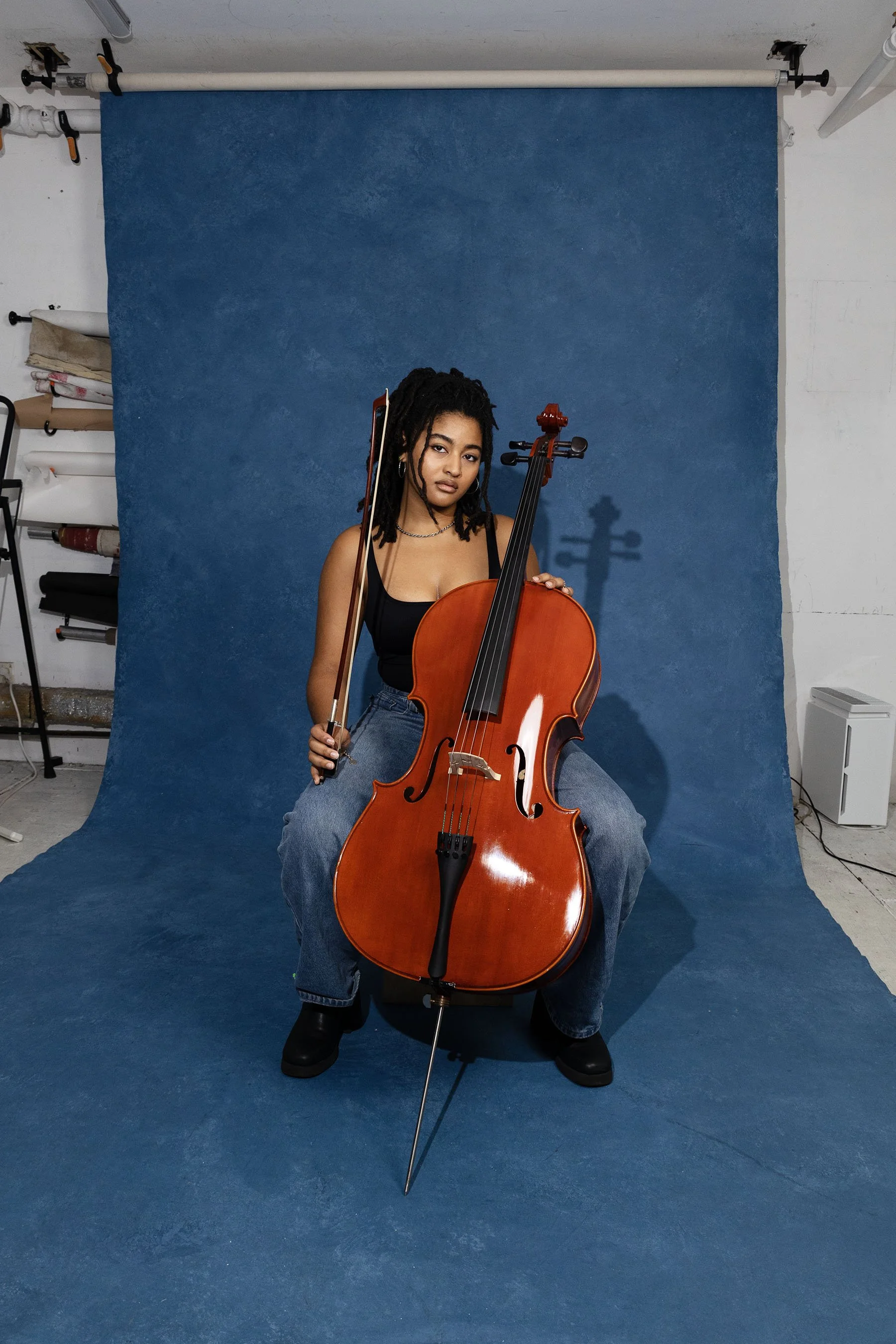Woman with dreadlocks sitting on the floor holding a cello, in a photography studio with a blue backdrop and various equipment on the side.