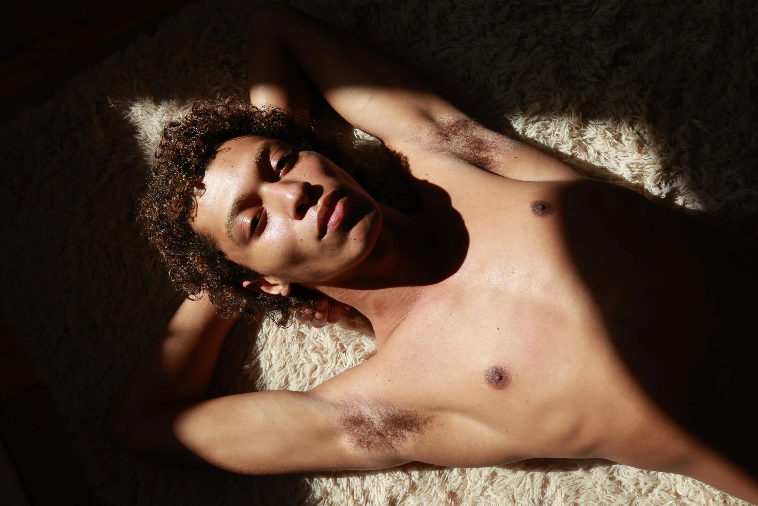 A woman with curly hair lying on a soft, textured surface in sunlight, with her arms extended above her head.