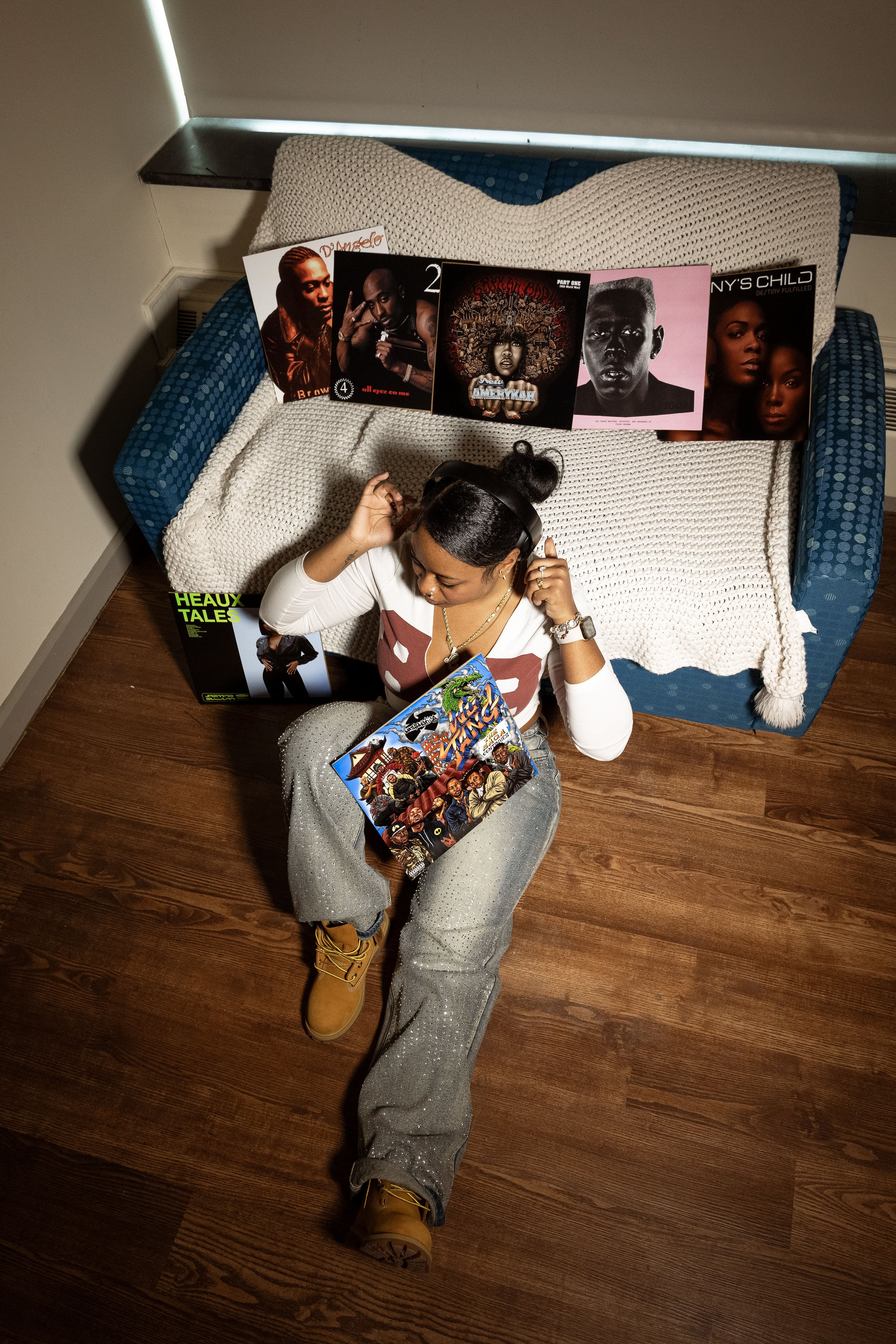 A woman lying on a wooden floor with her head resting on a beige blanket and wearing yellow boots, glasses, a white and red shirt, and gray pants. She has a magazine on her lap and several music and art posters spread on the bed behind her.
