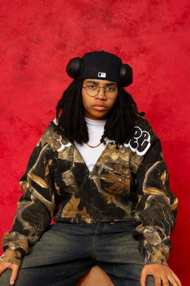 A young woman with glasses and dreadlocks wearing a black hat with headphones, a camouflage jacket, and sitting against a red textured background.