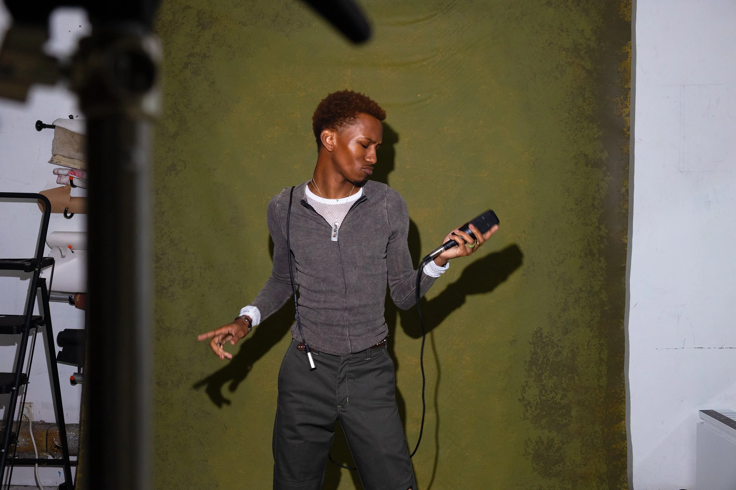 A young man with curly hair wearing a gray jacket, holding a microphone, appears to be dancing or performing against a green wall.