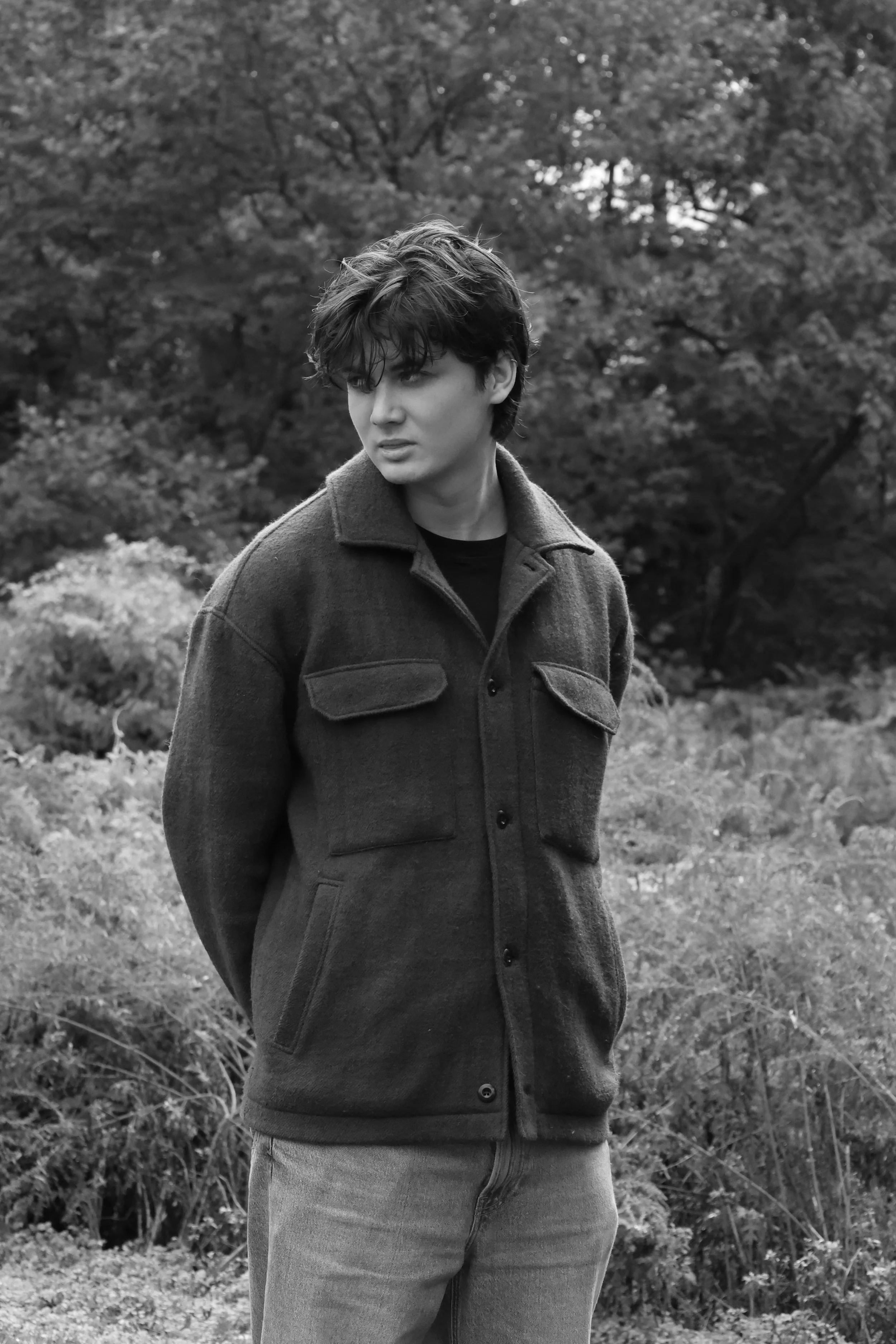 A person with short, curly hair wearing a dark jacket and light-colored pants standing outdoors in front of trees and grass.