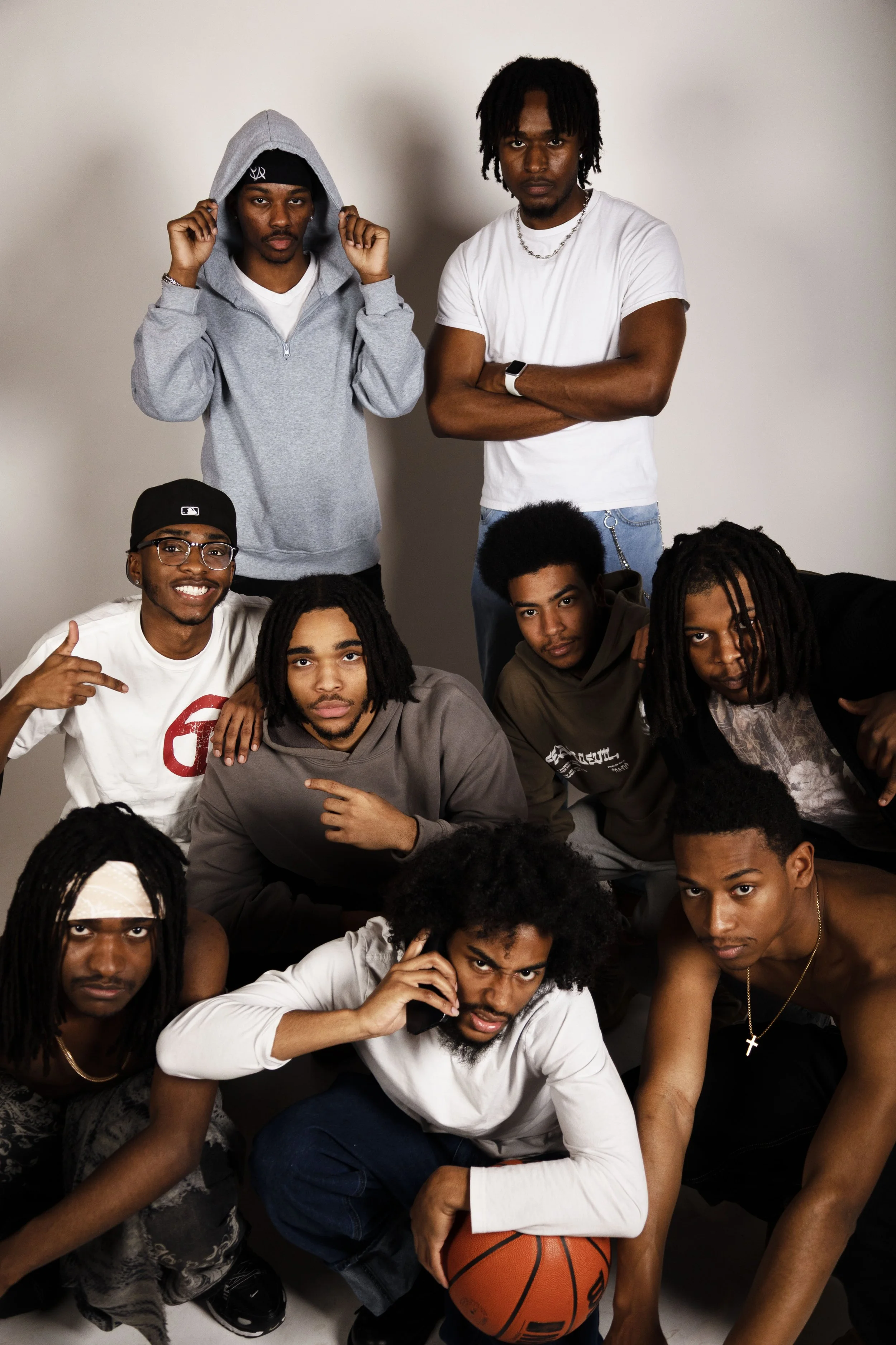 A group of young men poses together, some standing and others sitting, against a plain wall. They have various hairstyles, including dreadlocks and afro, and are dressed in casual clothing. One man is holding a basketball.