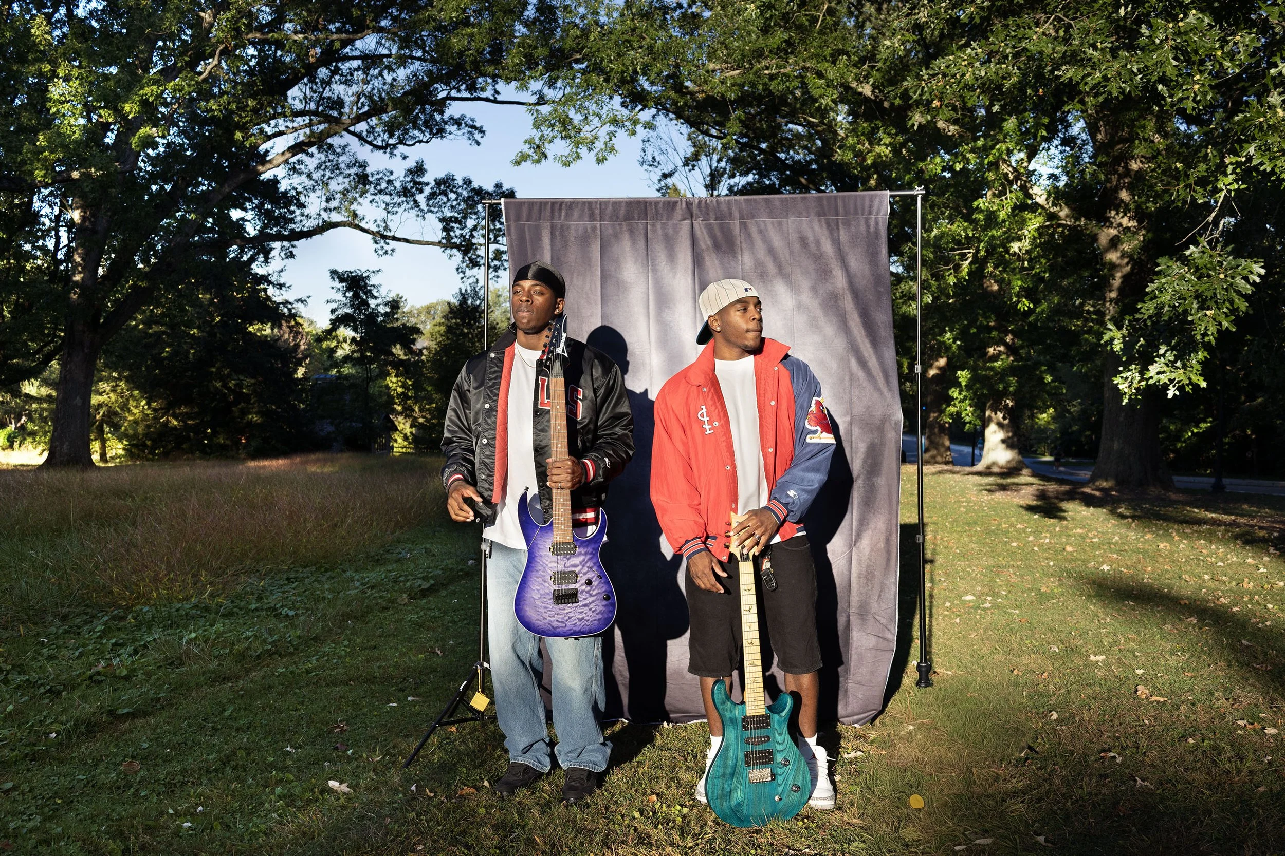 Two young men standing outdoors in front of a gray backdrop surrounded by trees, each holding a guitar, one with a purple electric guitar and the other with a turquoise electric guitar, dressed in casual athletic wear and jackets.