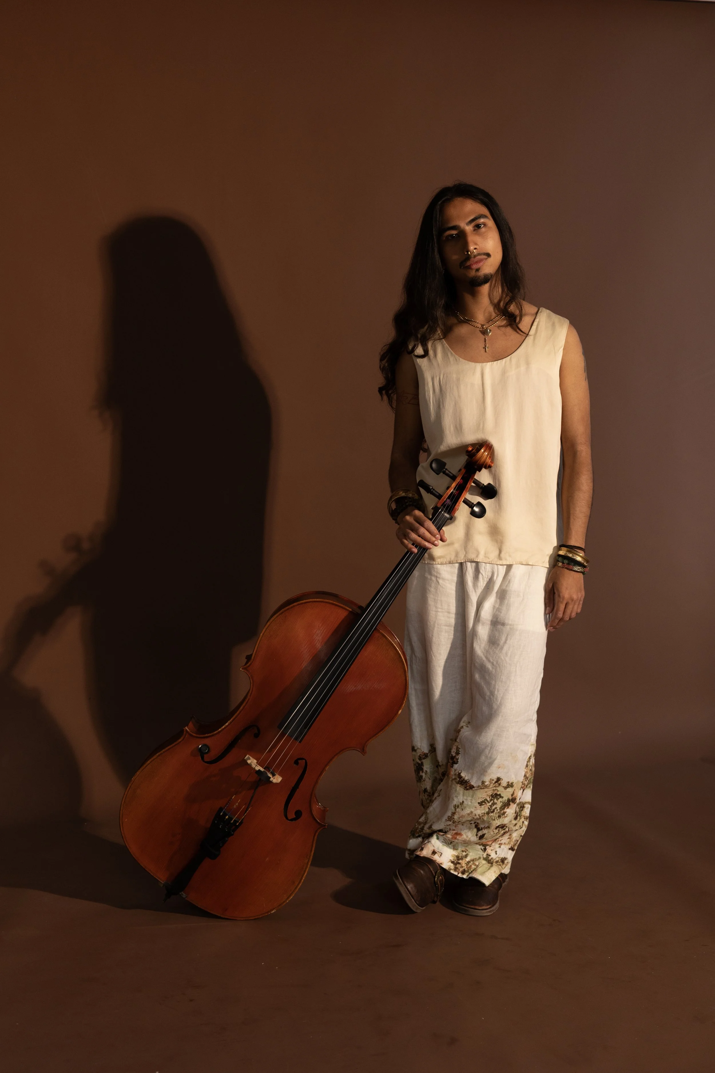 A person with long dark hair, wearing a beige sleeveless top and white patterned pants, holding a cello, standing against a plain brown background with a shadow cast on the wall.