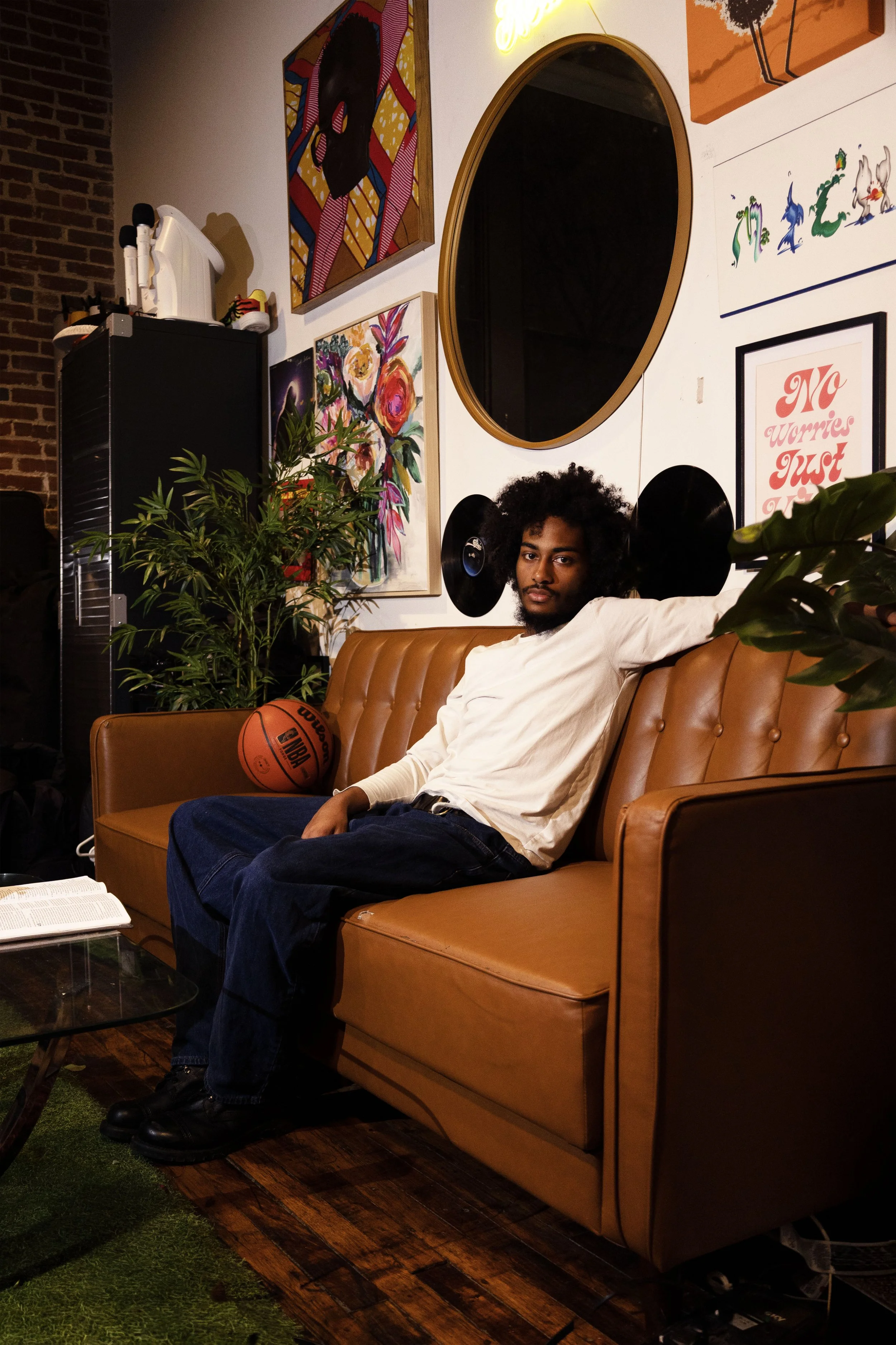 A young man with a curly afro hairstyle and a beard sitting on a tan leather couch. He is wearing a white shirt and dark jeans, with a basketball resting beside him. The background features colorful paintings, a large oval mirror, vinyl records on th