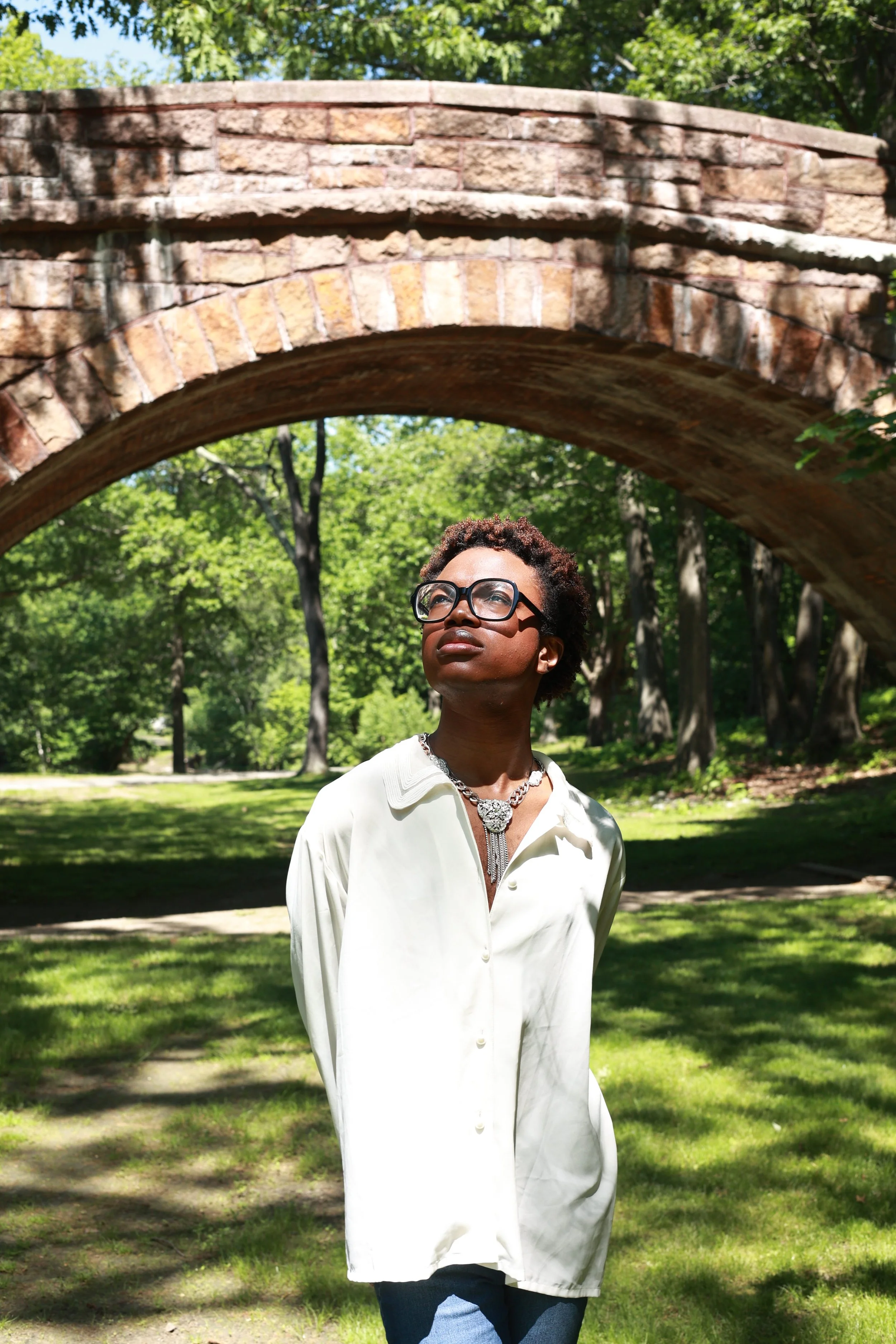 A woman with short, curly hair and glasses standing outdoors under a large brick archway, with trees and green grass in the background.