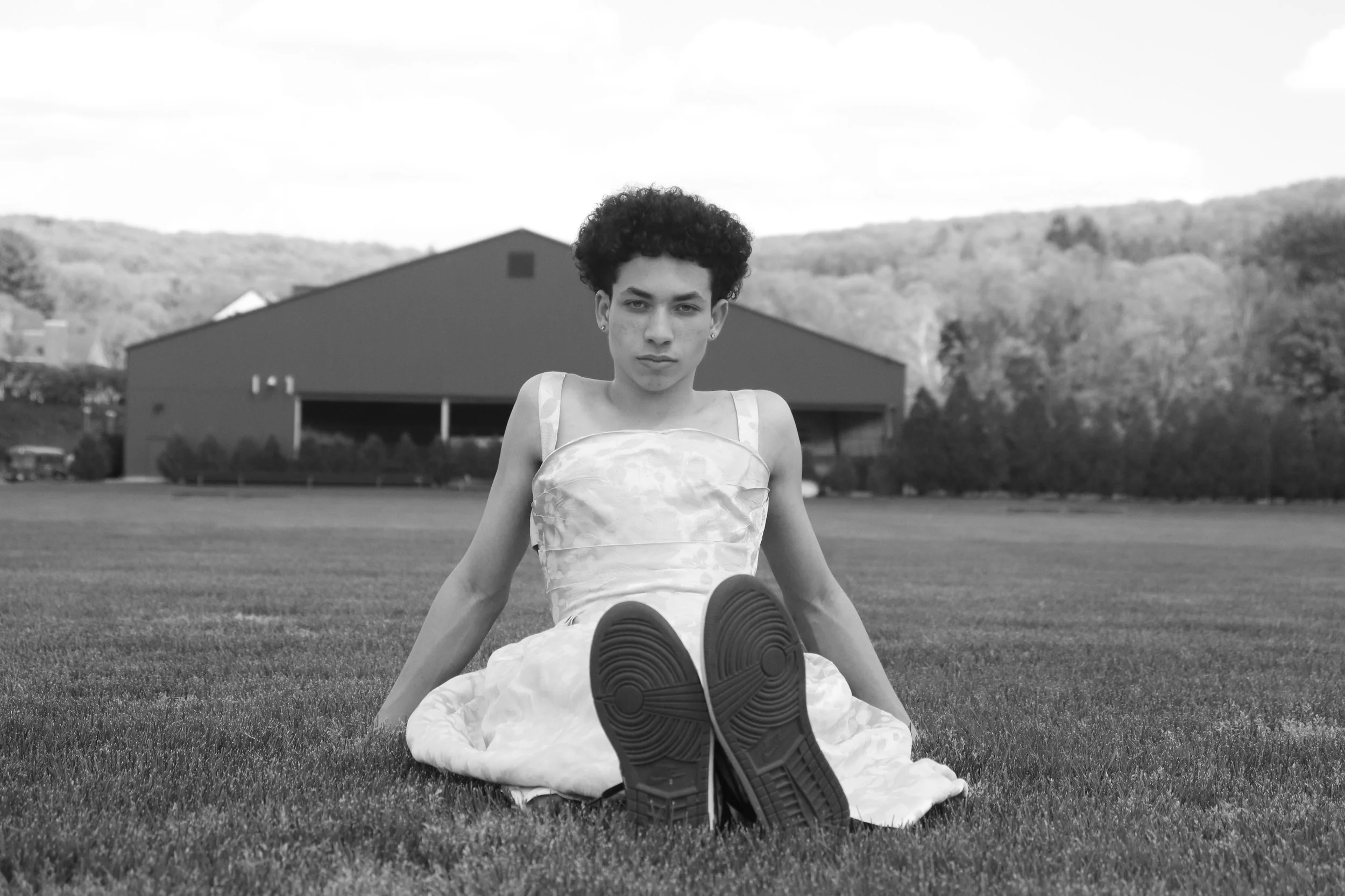 A young person with short curly hair sitting on the grass in a dress, facing the camera. They are outdoors with a house and trees in the background, and appear to be relaxing.
