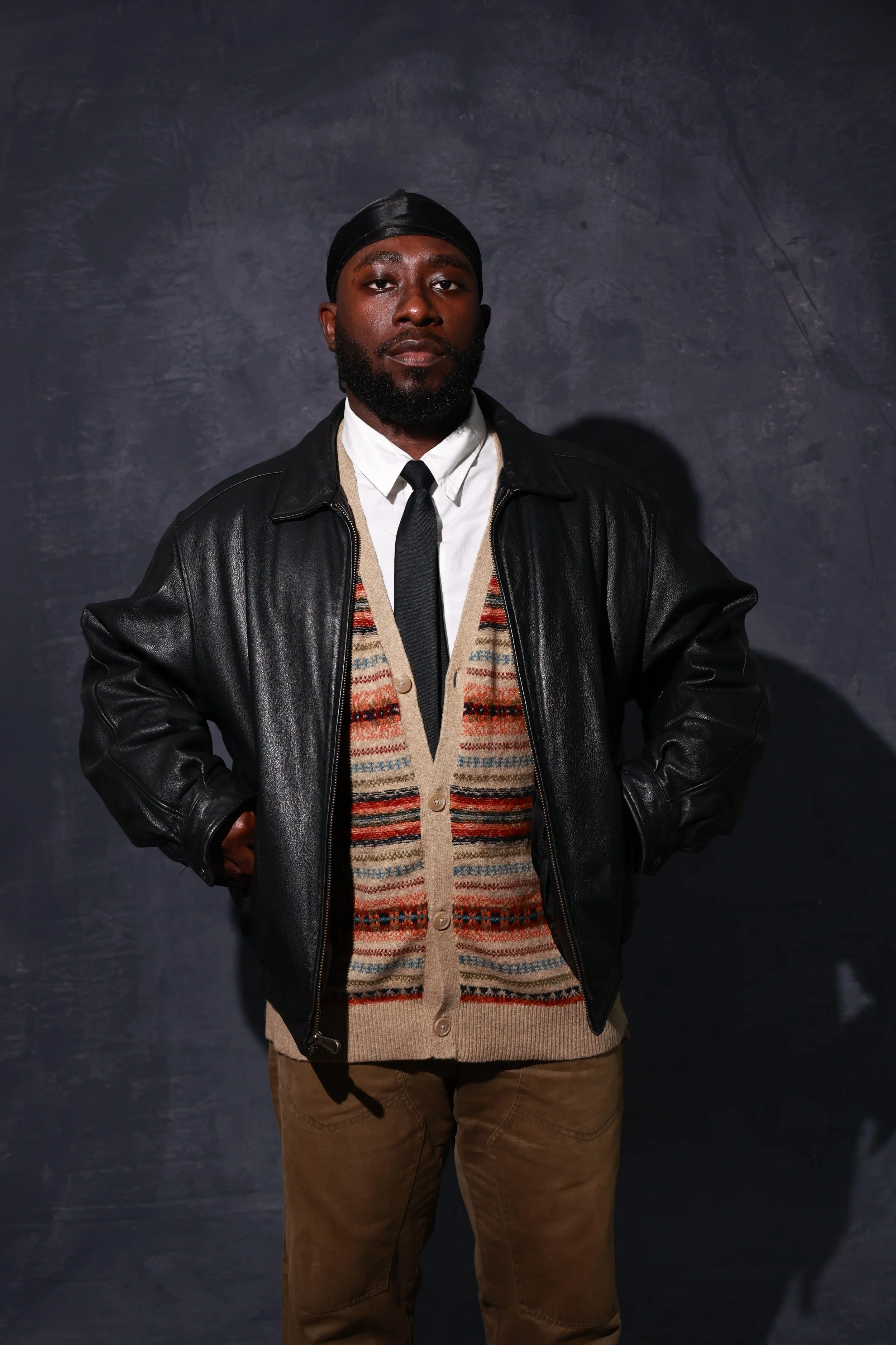 portrait of a man wearing a black leather jacket, white shirt, black tie, and beige sweater with colorful patterns, standing against a dark textured background.