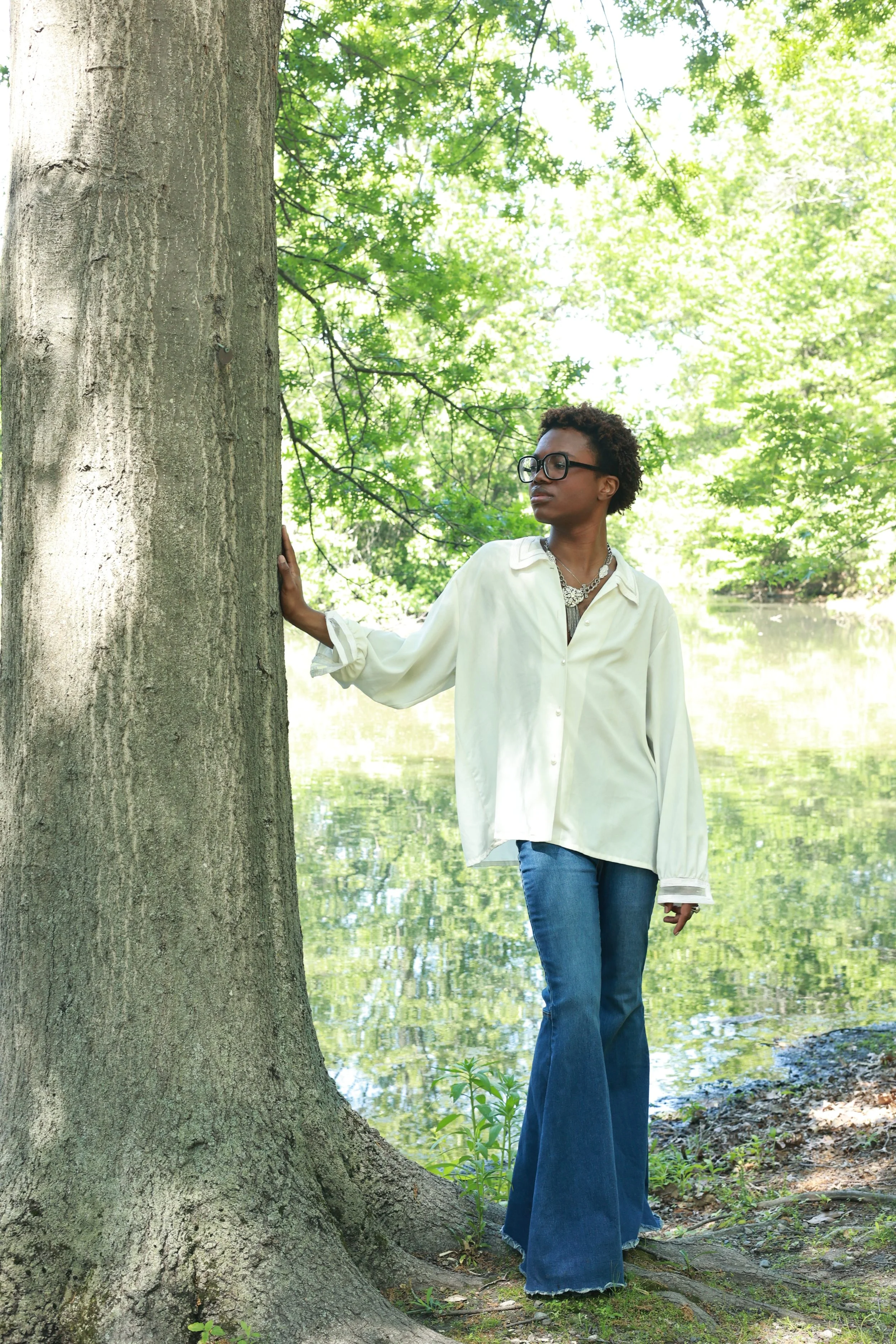 A woman with short curly hair, glasses, wearing a cream-colored blouse and high-waisted wide-leg jeans, stands near a large tree by a body of water in a lush green park during daytime. She has one hand on the tree trunk and looks to her left.