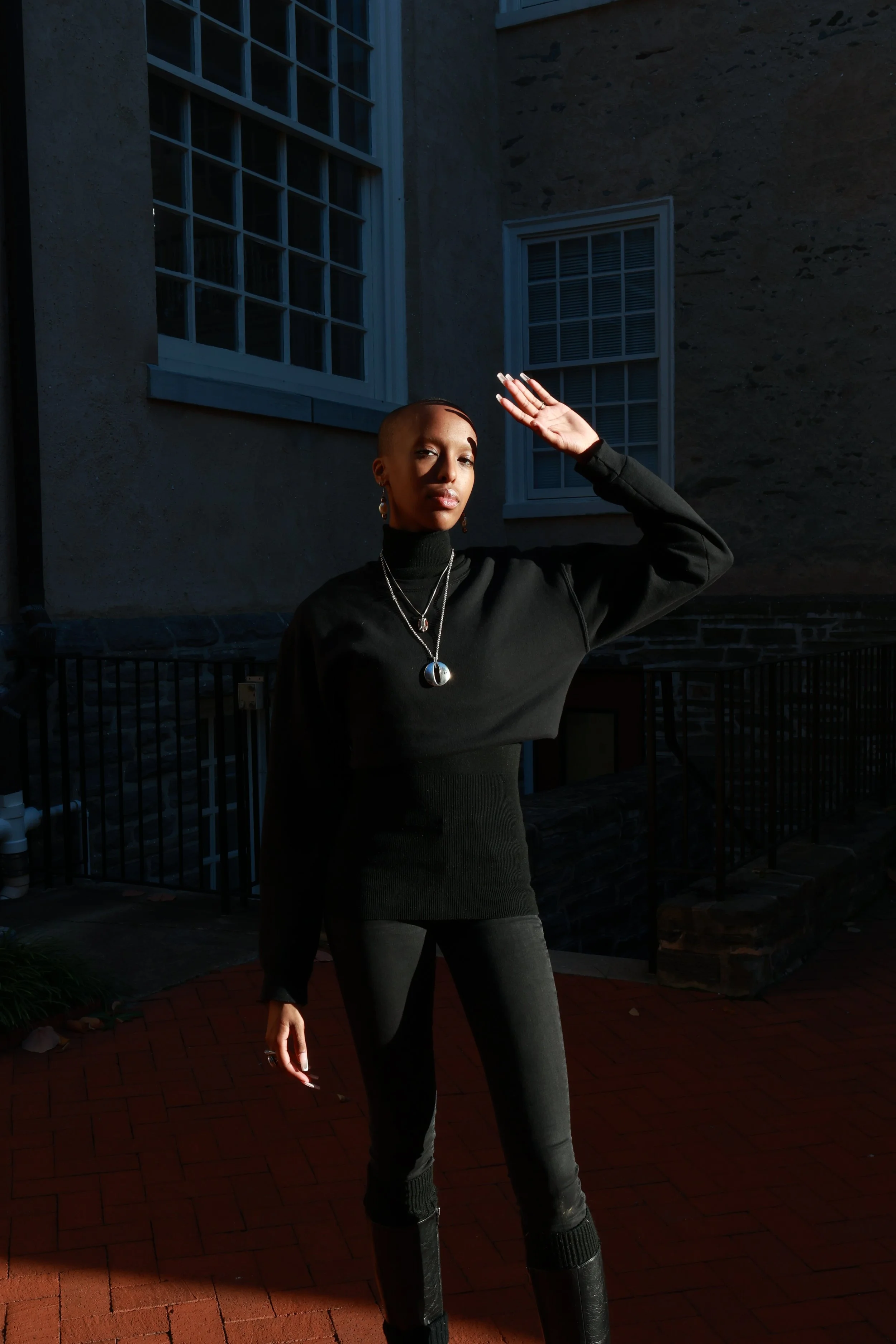 A woman wearing a black turtleneck sweater and black pants standing outdoors on a brick patio with her hand raised to her forehead, near two windows in a building with textured walls.