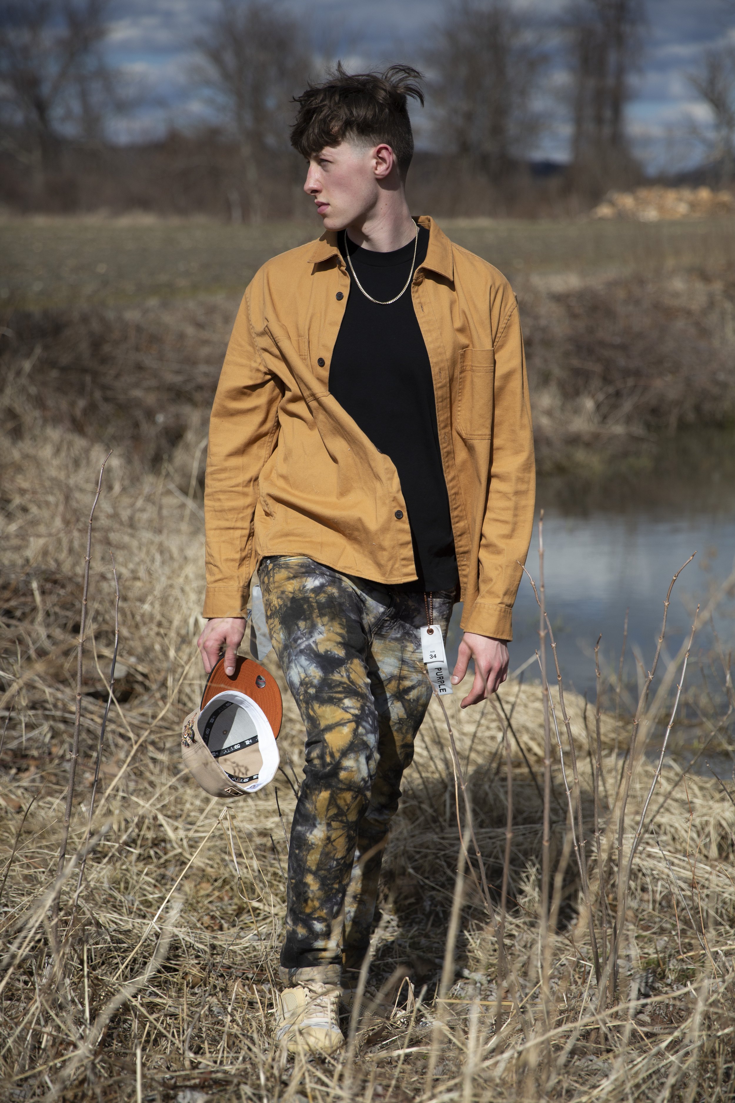 A young man walking through a dry, grassy outdoor area during daytime, holding a beige cap and wearing camouflage pants, a black shirt, a tan jacket, and a silver necklace, with a lake or pond in the background and a partly cloudy sky.