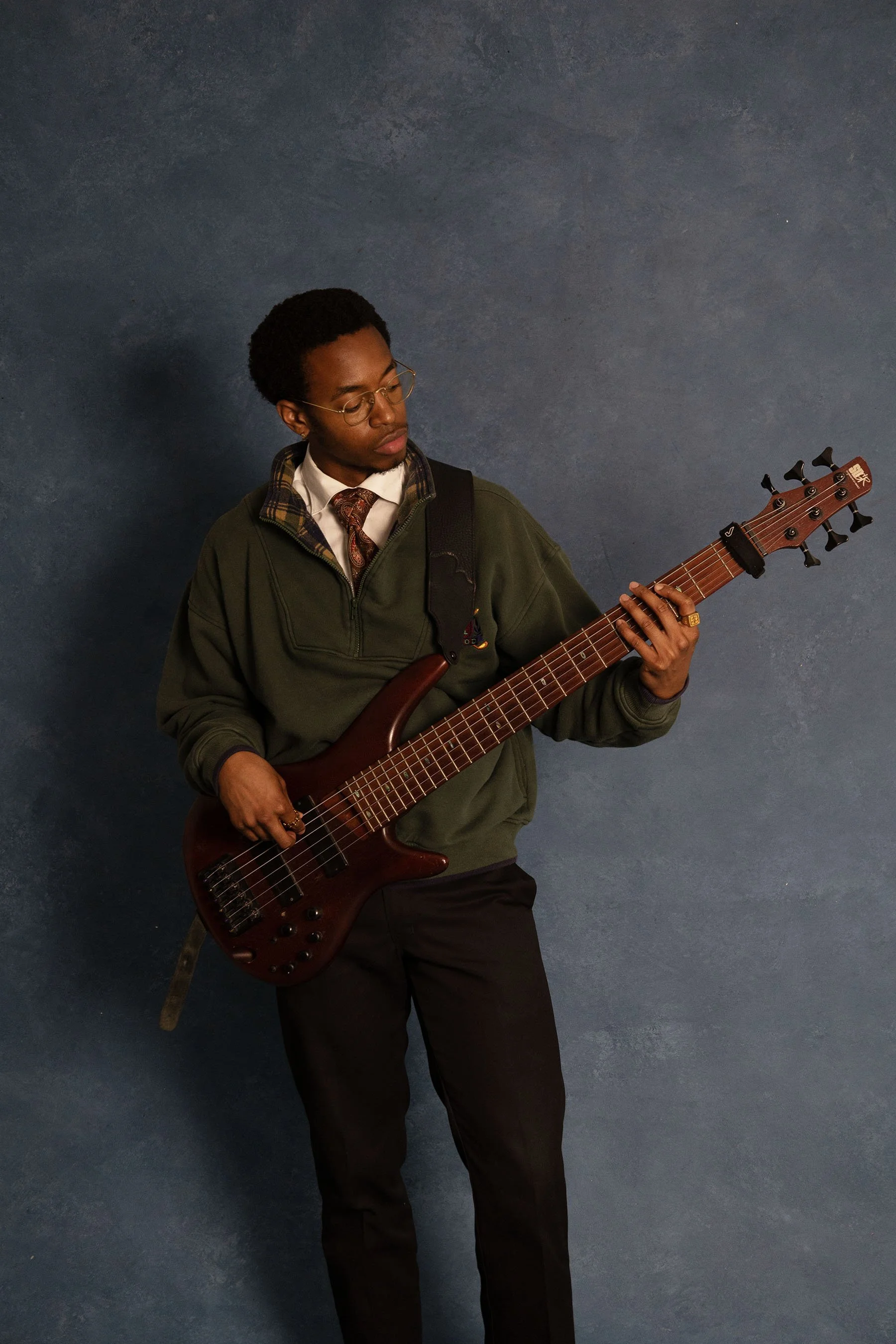 A young man with glasses and an afro hairstyle playing an electric guitar against a blue textured wall. He is wearing a green jacket, white shirt, and a patterned tie.