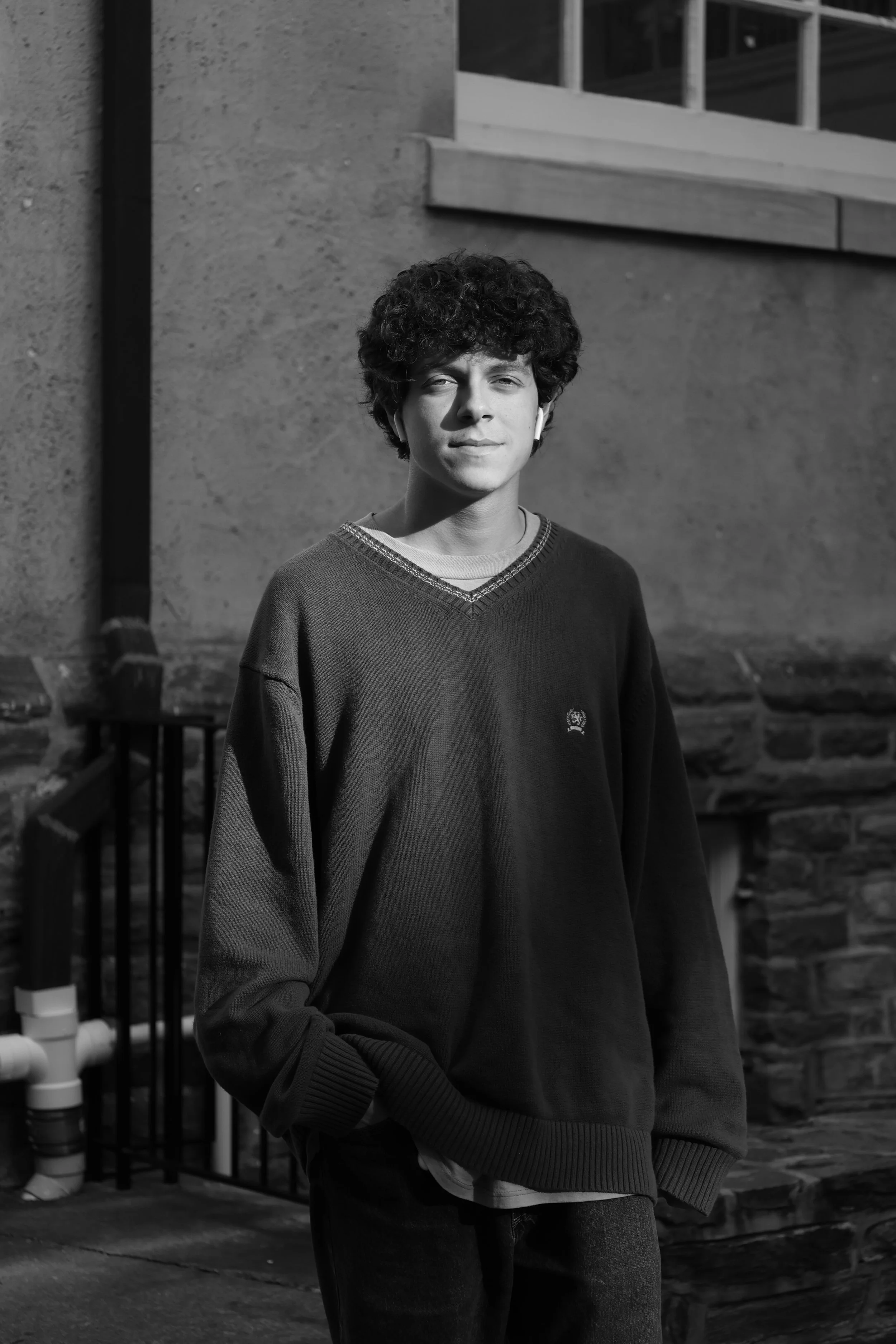 A young person with curly hair wearing a dark sweatshirt and jeans, standing outdoors near a brick wall, with sunlight on their face.