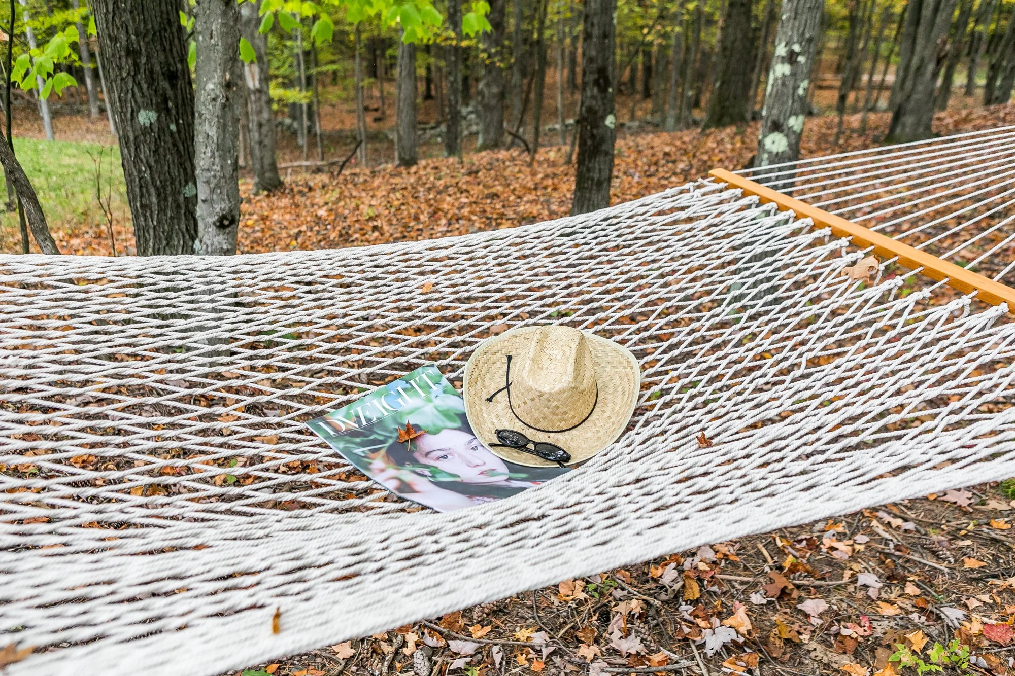 A hammock strung between trees in a forest with fallen leaves on the ground. On the hammock, there is a magazine, a straw hat and a pair of sunglasses indicating a relaxing setting.