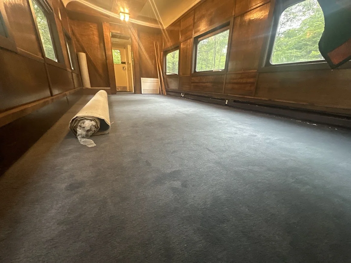 A finished train car lounge with wooden walls and large windows and freshly installed blue carpet.