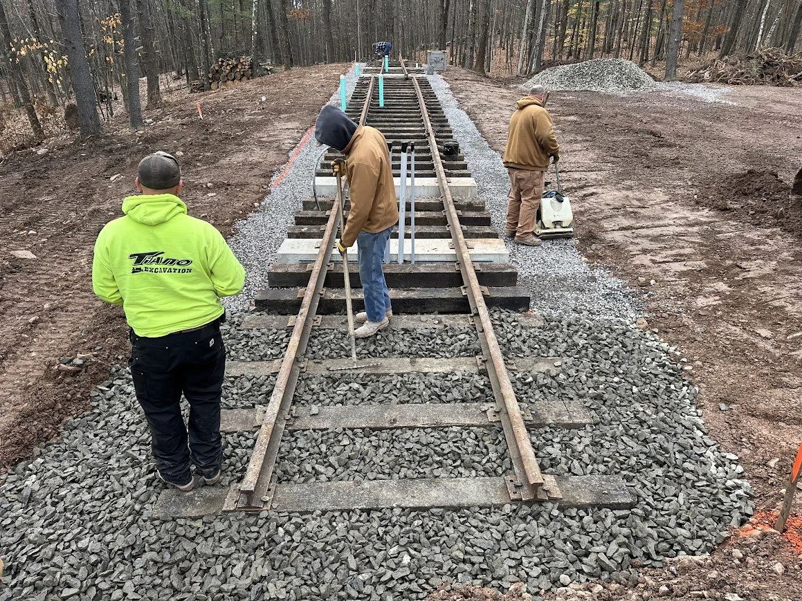 Workers repairing and constructing a railway track in a wooded area, with gravel surrounding the tracks and construction equipment nearby.