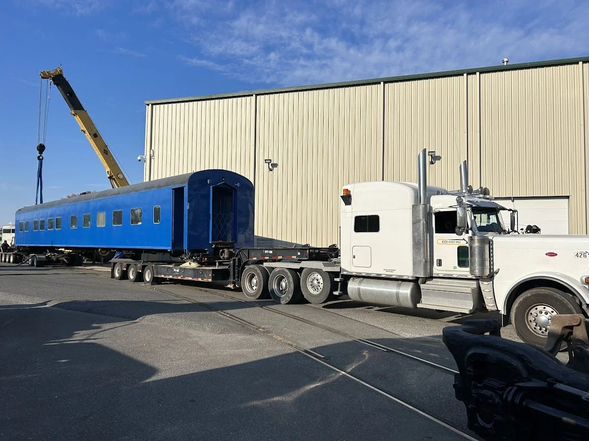 A white semi-truck with an attached flatbed trailer carrying a large blue train car is parked at a rail yard with a crane lifting the train car into place.