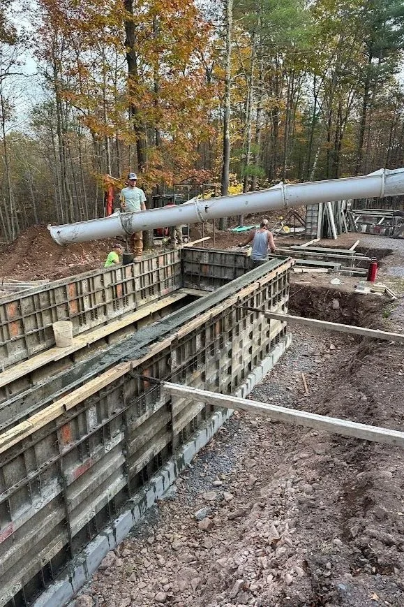 Construction workers are building a concrete form with wooden and metal framing, with a large pipe overhead, in a wooded area with trees and autumn leaves.