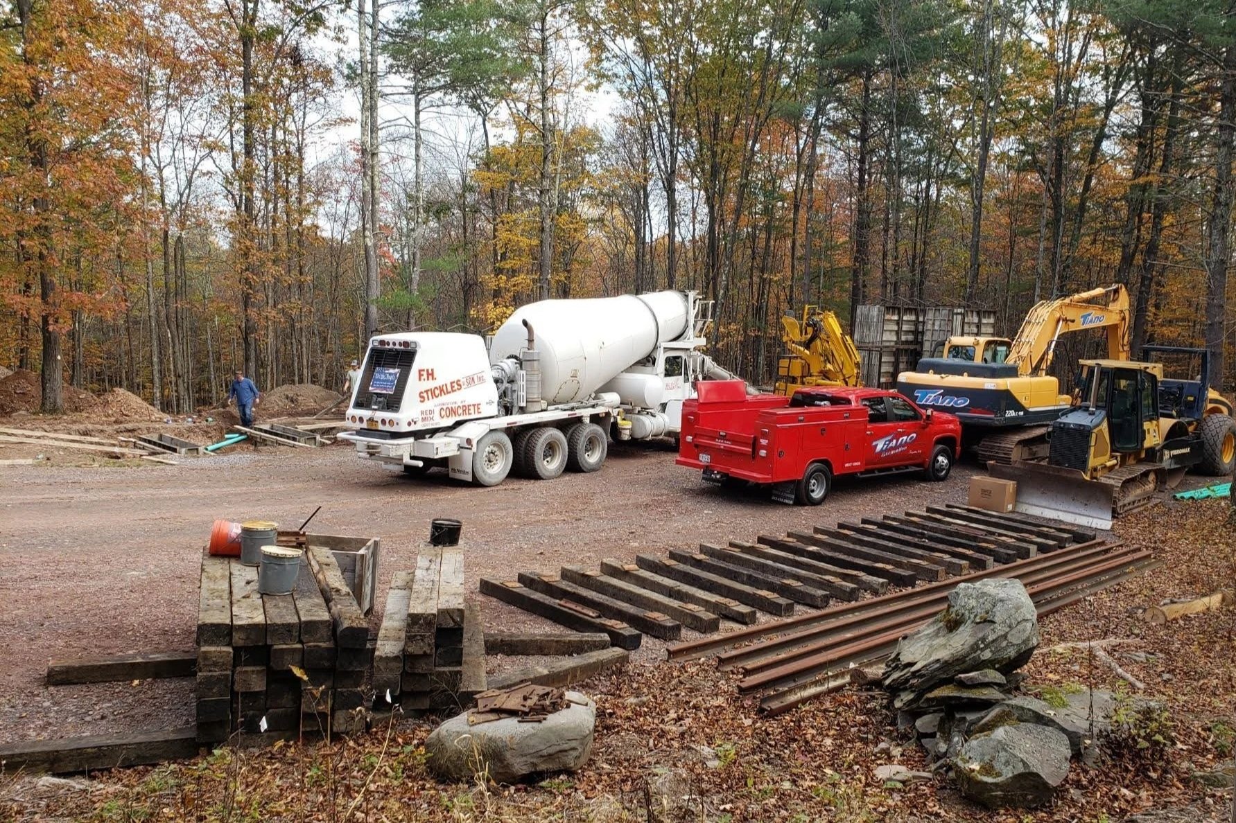 Construction site with trucks, excavators, and machinery in a wooded area with fall foliage.