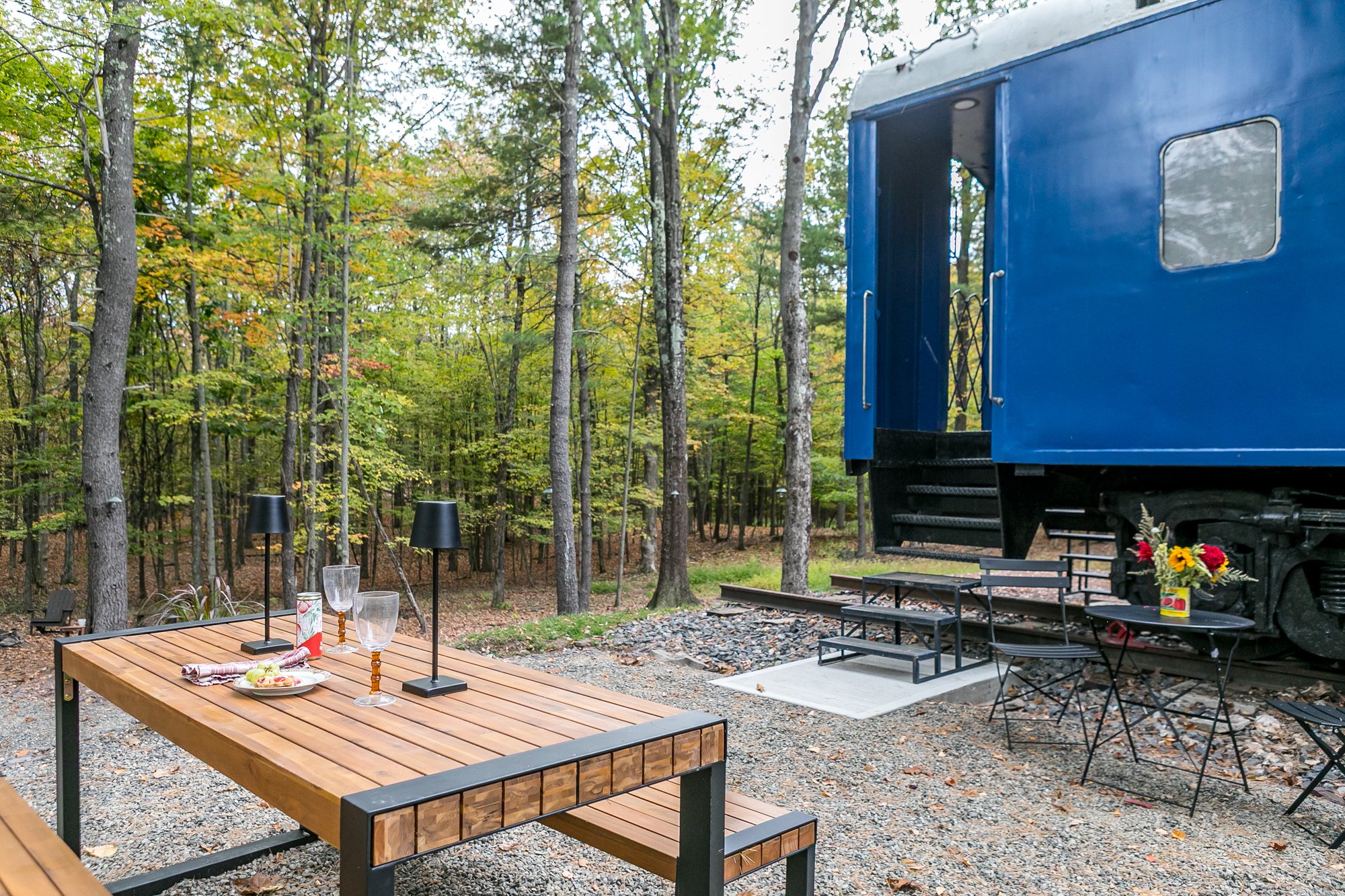 An outdoor dining area in a woodland setting with a wooden table, lamps, glassware, and plates, with a blue train car and trees in the background. Also available are a grill and a patio set.