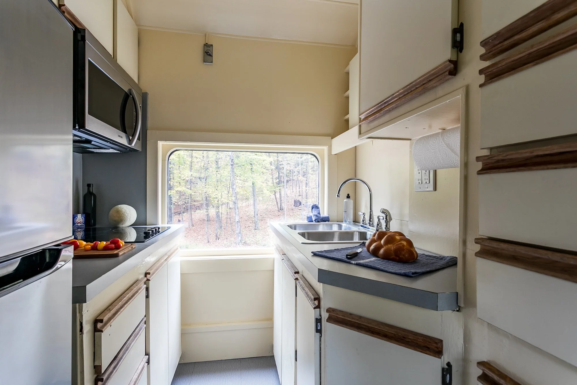 A cozy kitchen filled with natural light, featuring cream-colored walls, white cabinets, and wooden trim. A window overlooks a wooded area. There is a double sink, microwave, stovetop and refrigerator.