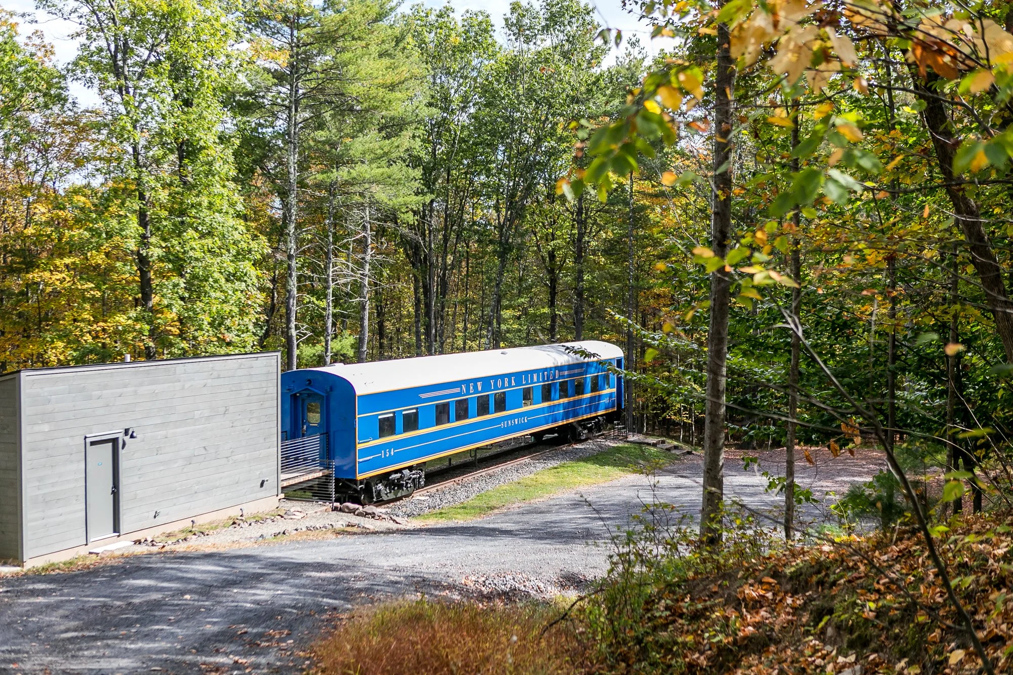 A vintage blue and yellow train car labeled ''New York Limited Sunswick" on tracks surrounded by green trees in a forest setting. A modern bathhouse and large driveway are also visible.