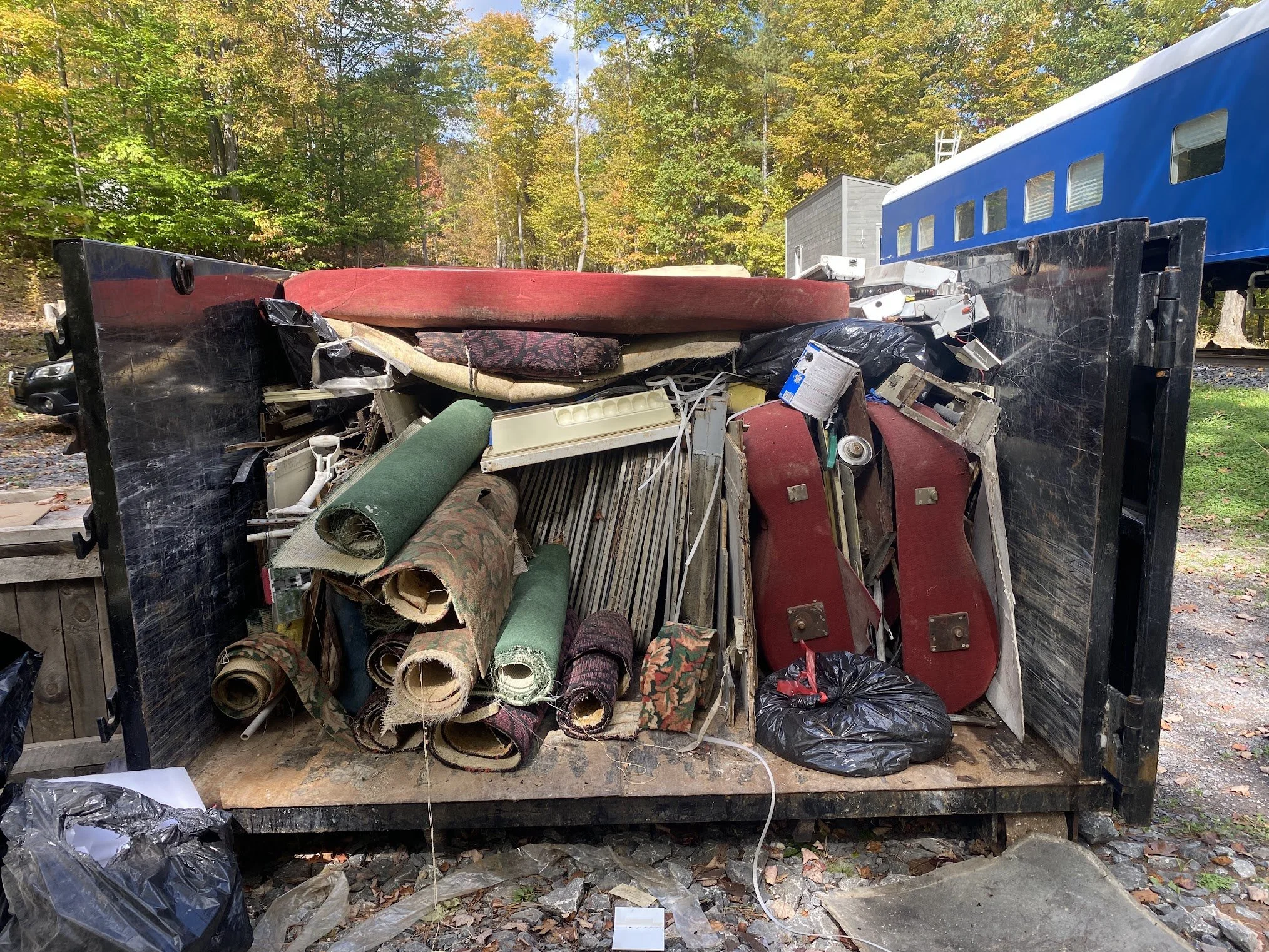 A dumpster filled with discarded furniture, carpets, and trash, parked outdoors with a background of colorful autumn trees.