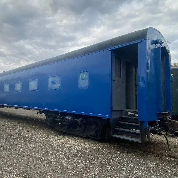 A long blue train car freshly painted with windows and a set of steps at the entrance, parked on railway tracks under a cloudy sky.